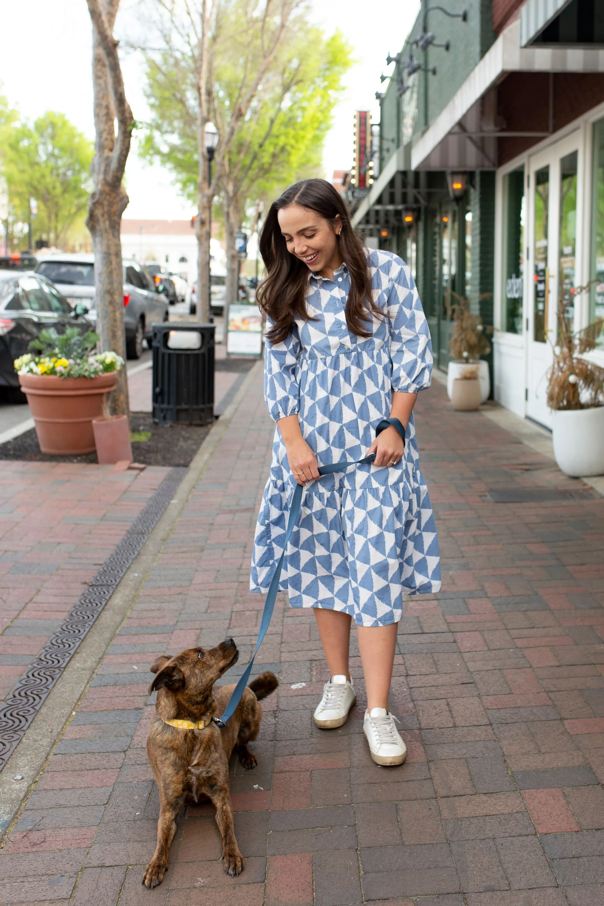 A young woman in a blue and white patterned dress walking her brown puppy on a city sidewalk with parked cars and storefronts in the background.