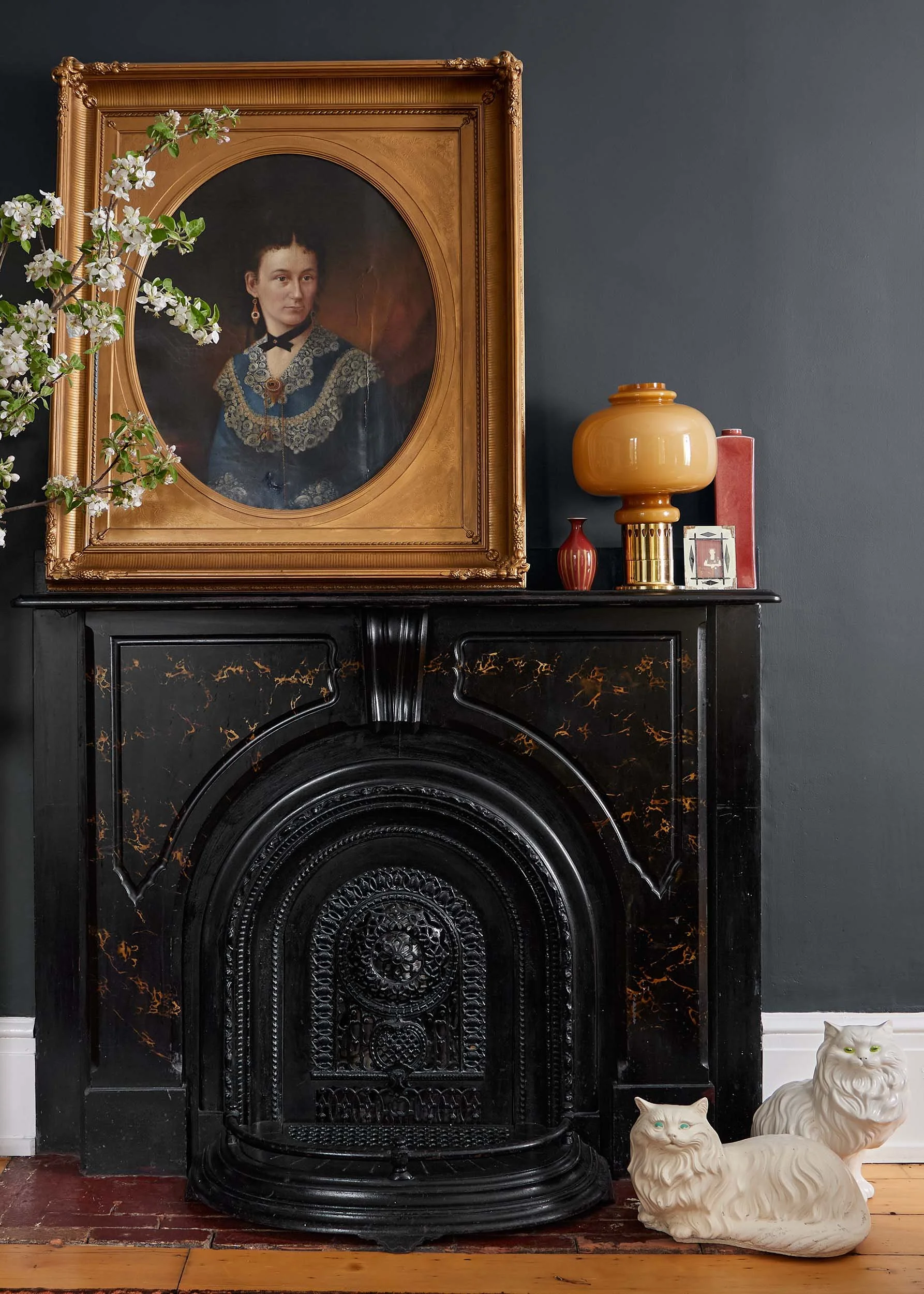 A vintage looking black marble fireplace with a detailed carved arch and ornate design. On top of the fireplace is a large, framed oval portrait of a woman in Victorian attire, with a dark background and a cracked surface on the painting. To the right of the fireplace are three decorative ceramic cats, with one in the foreground and two in the background. The wall behind is painted dark gray.