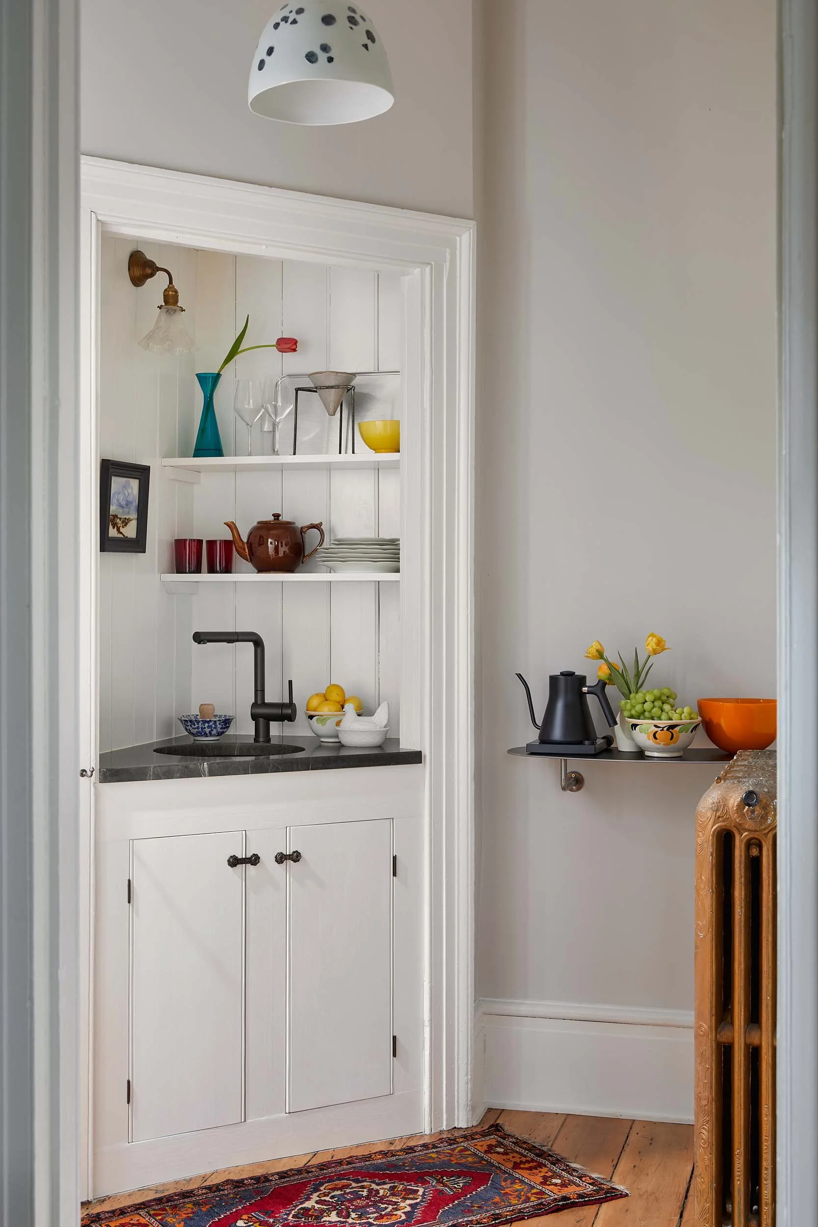 A cozy kitchen corner with a small black sink, a black kettle, bowls of lemons and grapes, a wooden pottery piece, and a white wall shelf with decorative glassware, a yellow bowl, a small framed photo, and a shelf with plates. French doors are partially visible, and there is a colorful patterned rug on hardwood floors.