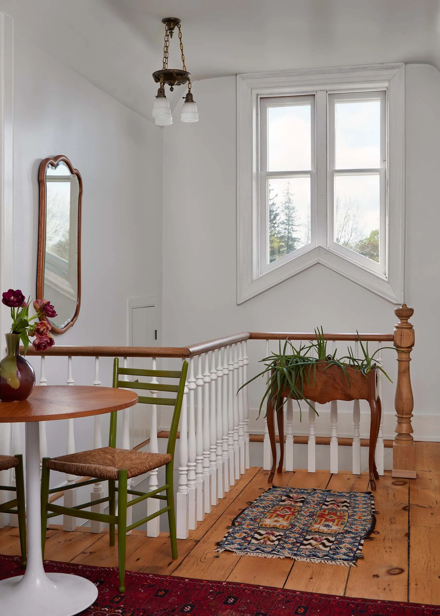 Interior view of a house with a staircase, wooden flooring, a small round table with a vase of flowers, a mirror on the wall, chairs, and a colorful patterned rug, with a large window and a chandelier hanging from the ceiling.