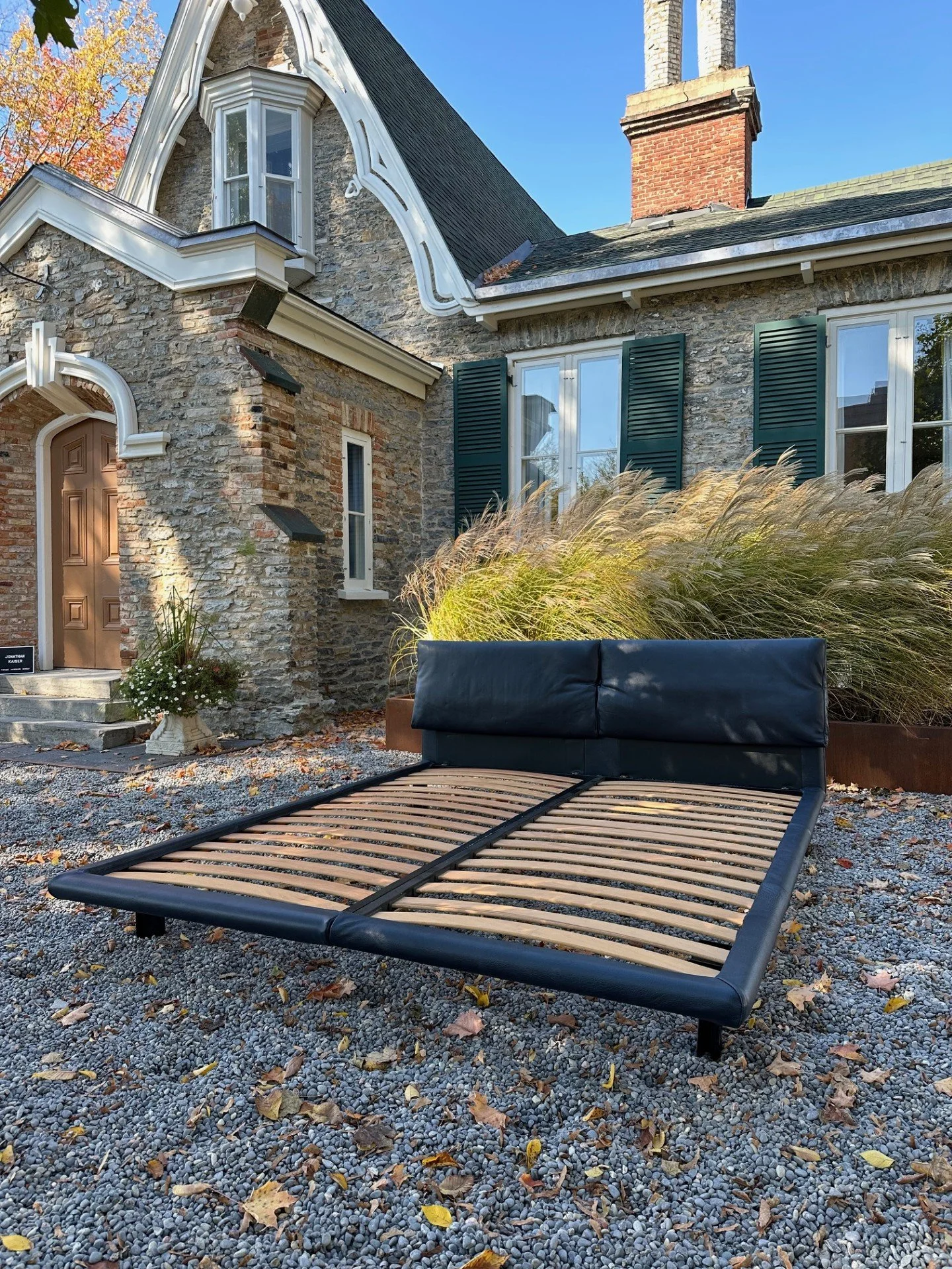 A black metal bed frame without a mattress is placed on a gravel surface in a backyard with fallen leaves. Behind it is a stone house with green shutters, white trim, and a pointed roof. The house has multiple windows and a brick chimney. There are ornamental grasses and a flower pot near the house entrance, and the sky above is clear blue with a few trees with autumn-colored leaves in the background.