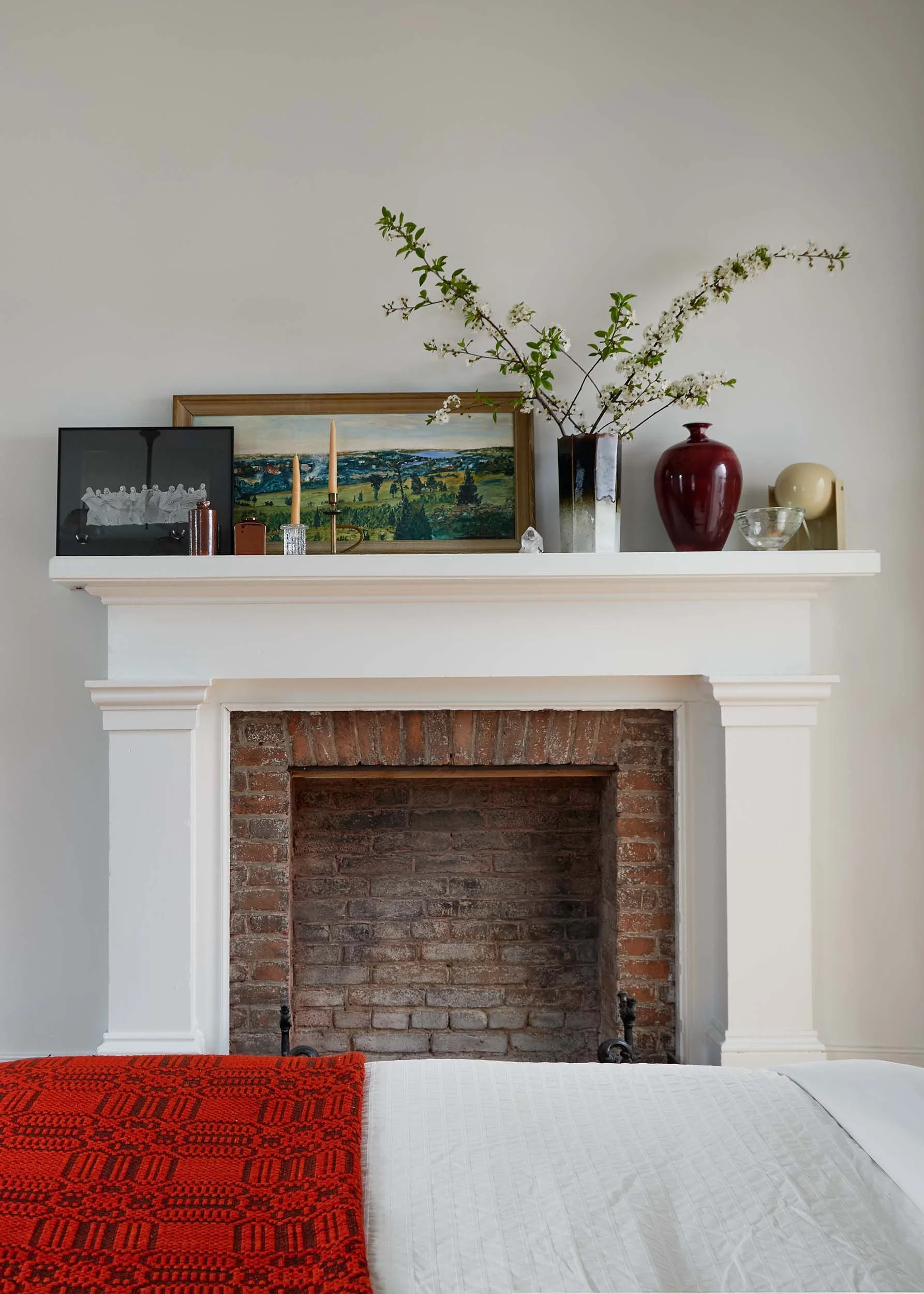 A bedroom with a fireplace and a white bed with a red blanket, decorated with a framed landscape painting, two candlesticks, a vase with white flowering branches, and decorative objects on the mantel.
