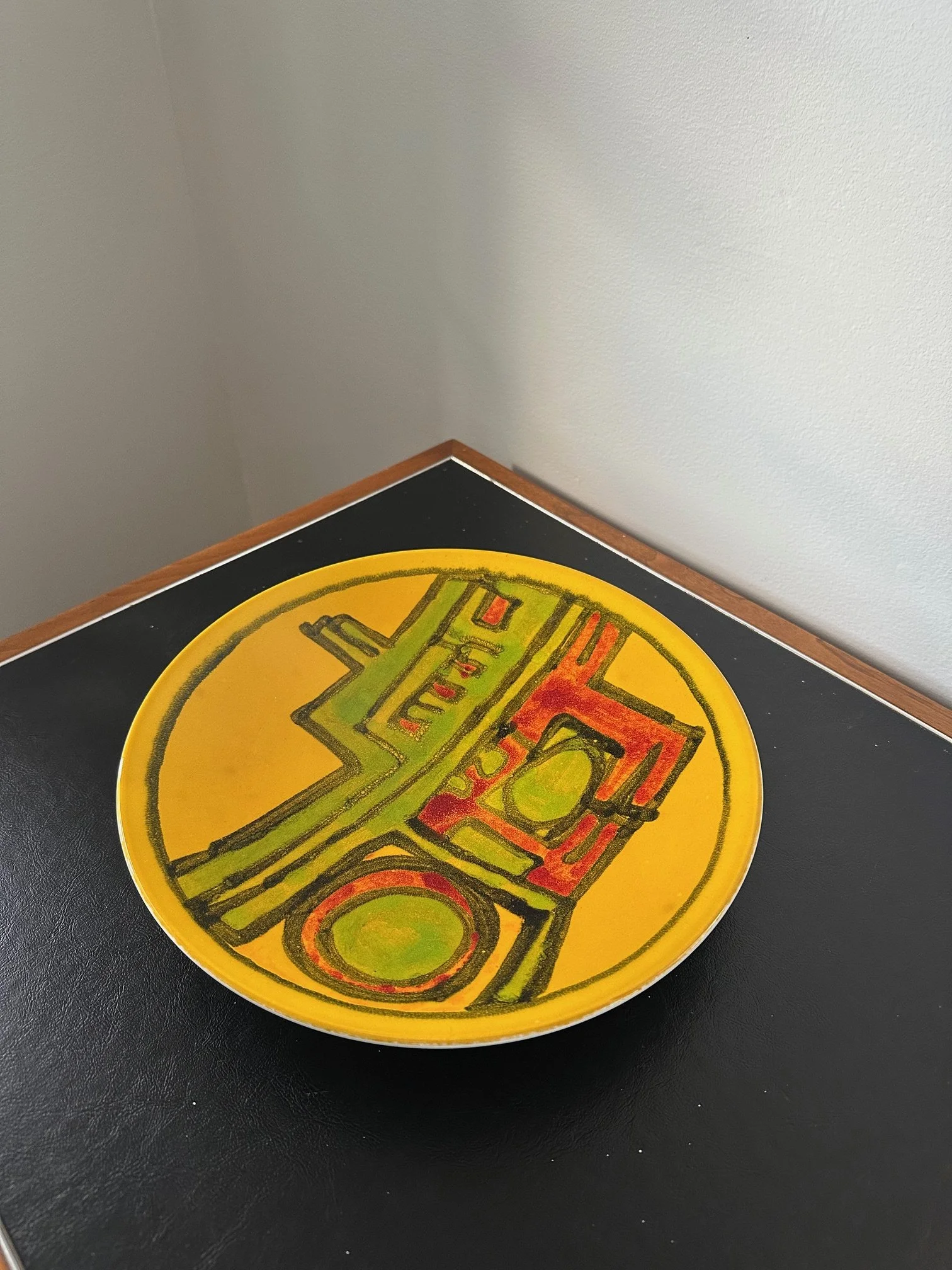 Colorful circular artwork with geometric shapes and patterns on a yellow background, placed on a black surface with a white wall in the background.