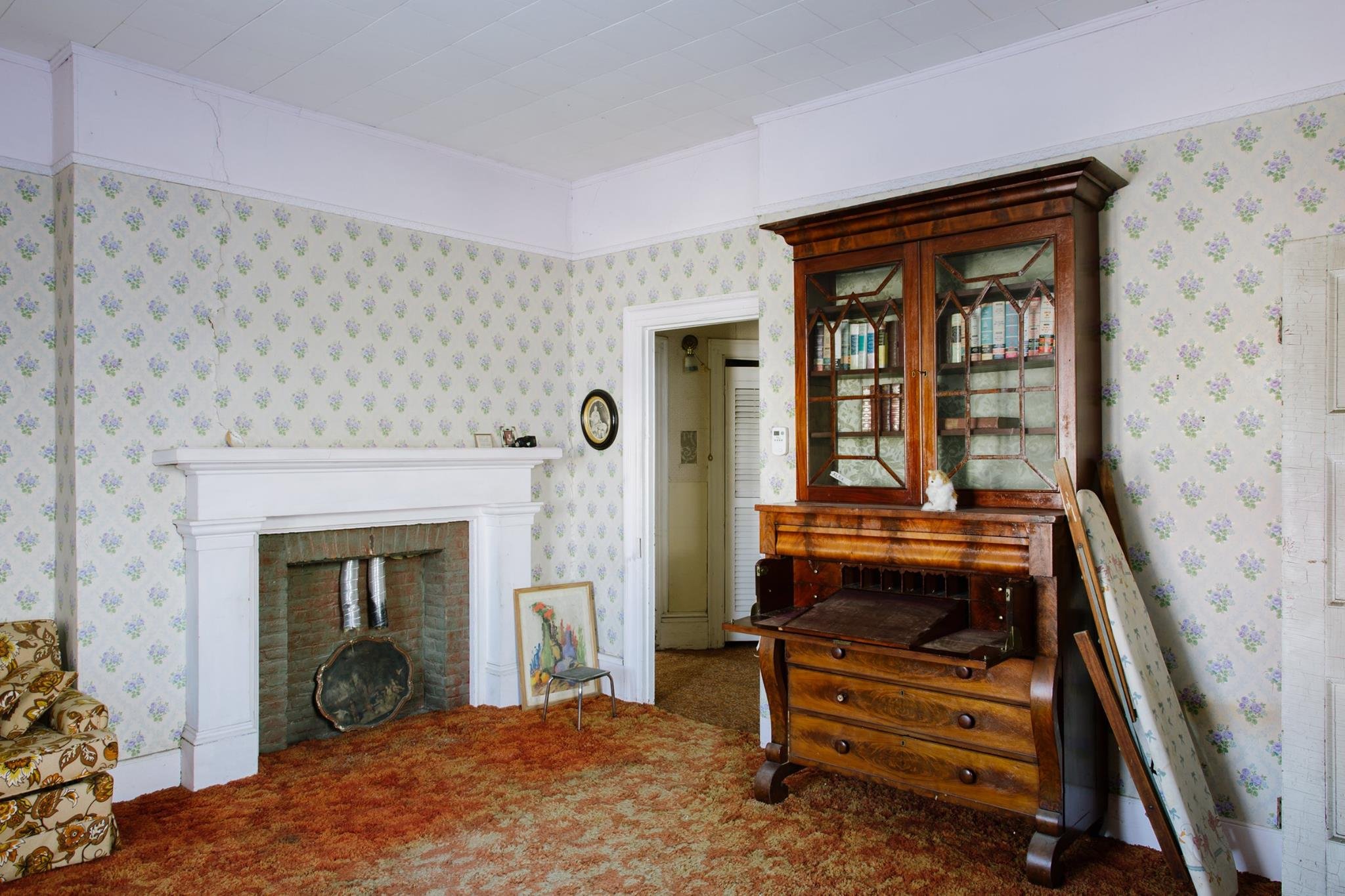 A vintage living room with floral wallpaper, a white fireplace, a brick interior, an ornate wooden china cabinet, and various small decorative items and artwork.