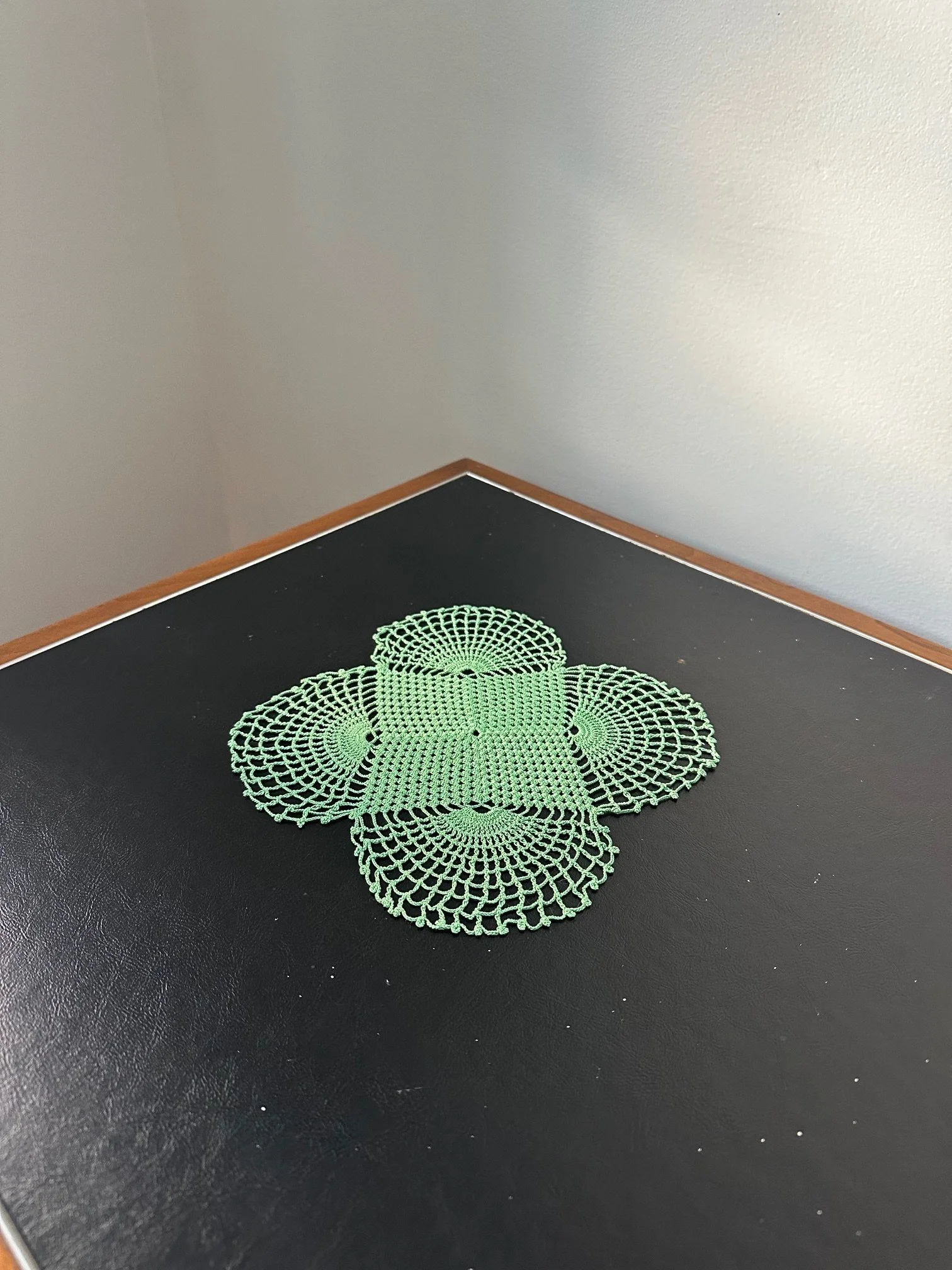 A green crocheted doily on a black surface with a gray wall and wooden trim in the background.