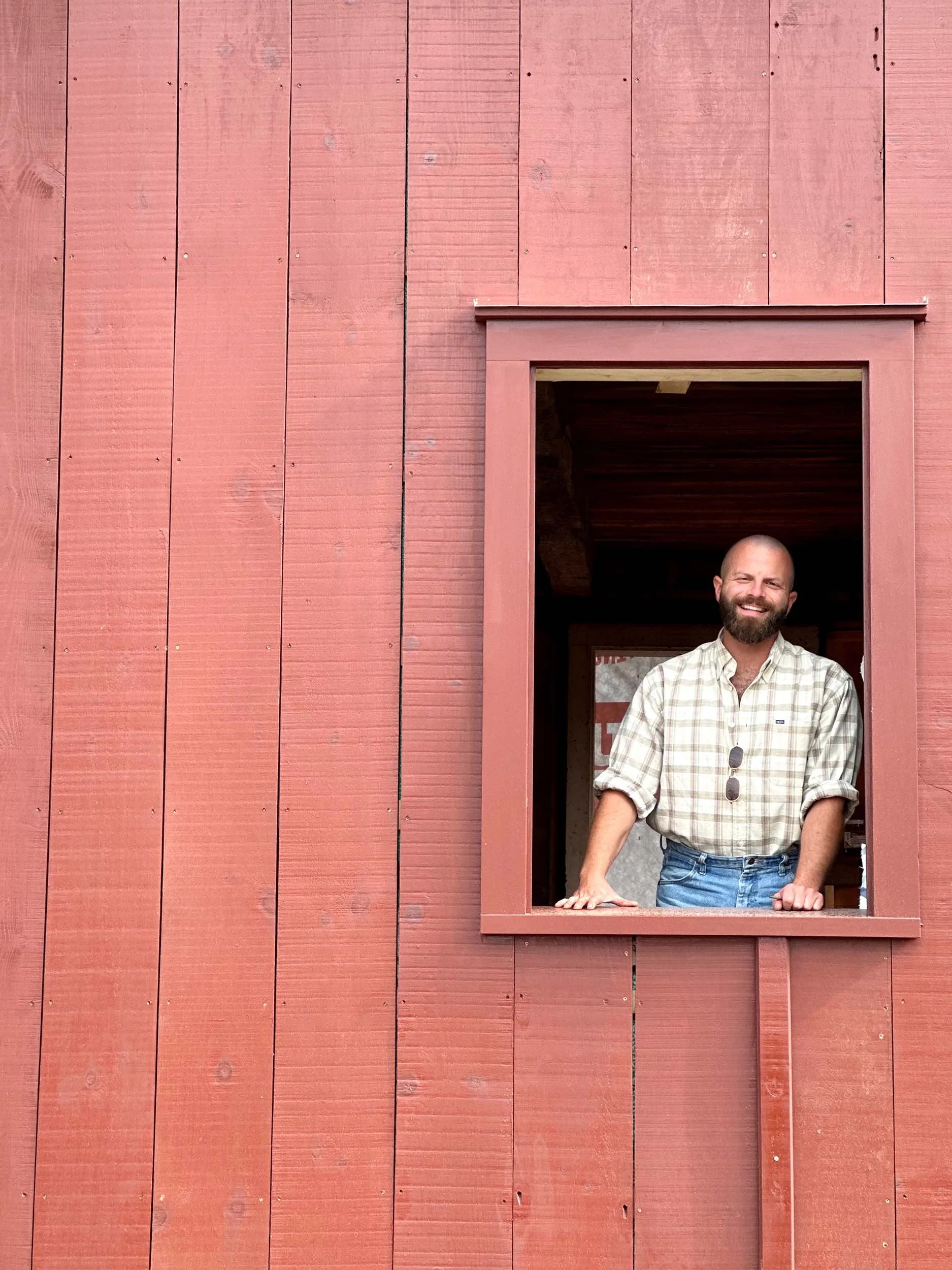 A man with a beard and short hair, wearing a plaid shirt and jeans, is standing in a window with his hands resting on the window sill, smiling at the camera. The building has red wooden siding.