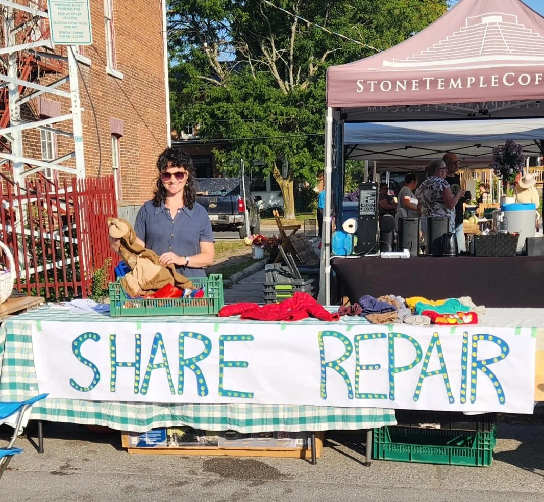Woman standing at a table with a sign that reads 'Share Repair,' selling clothing and textiles, at an outdoor market with tents and other booths, brick buildings, trees, and people in the background.