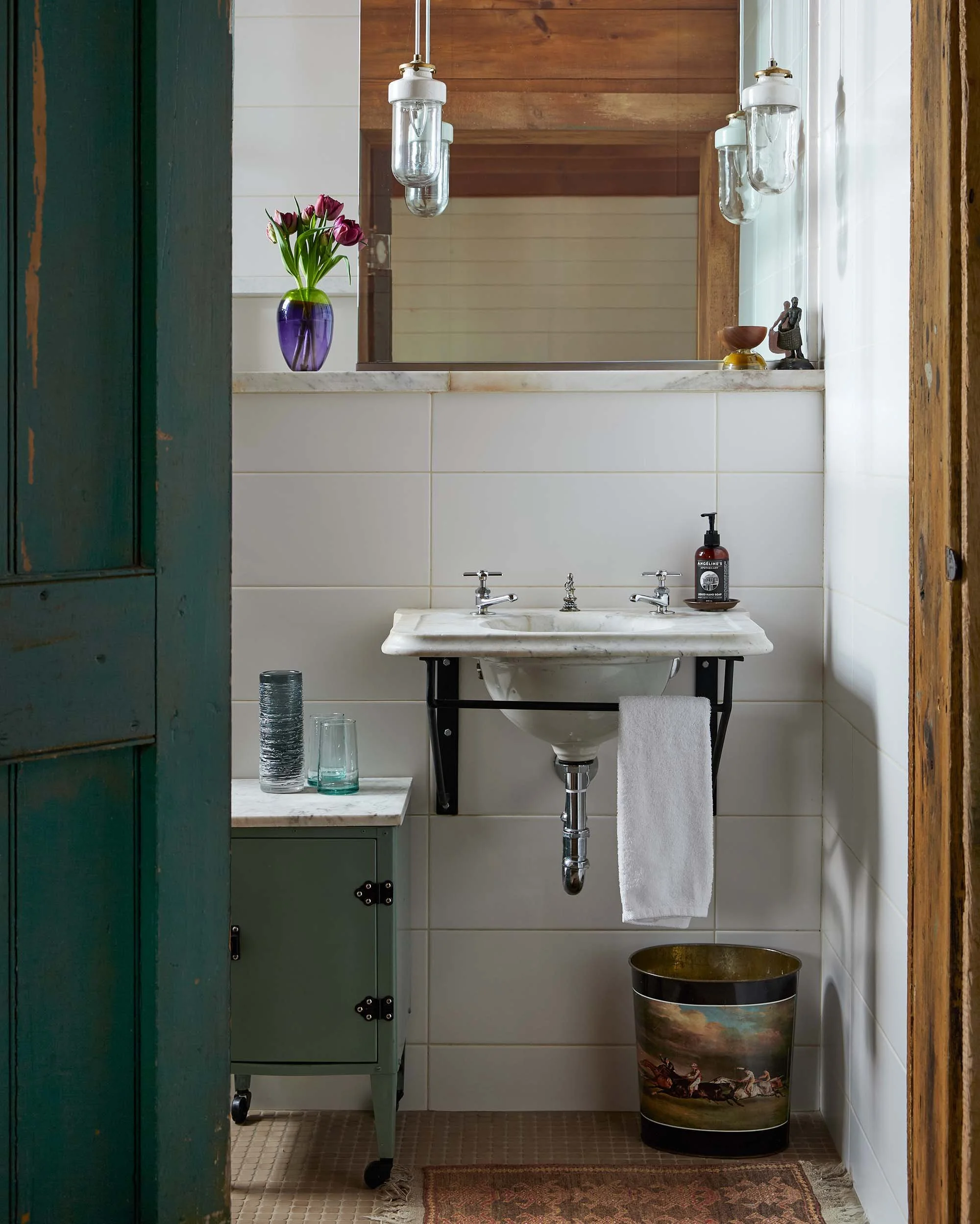 Bathroom with white tiled walls, a bust sculpture, a small vase, hanging glass jars, a double sink with faucets, a towel hanging below the sink, and a decorative bucket with a pastoral scene design, situated on a brown tiled floor with a rug.