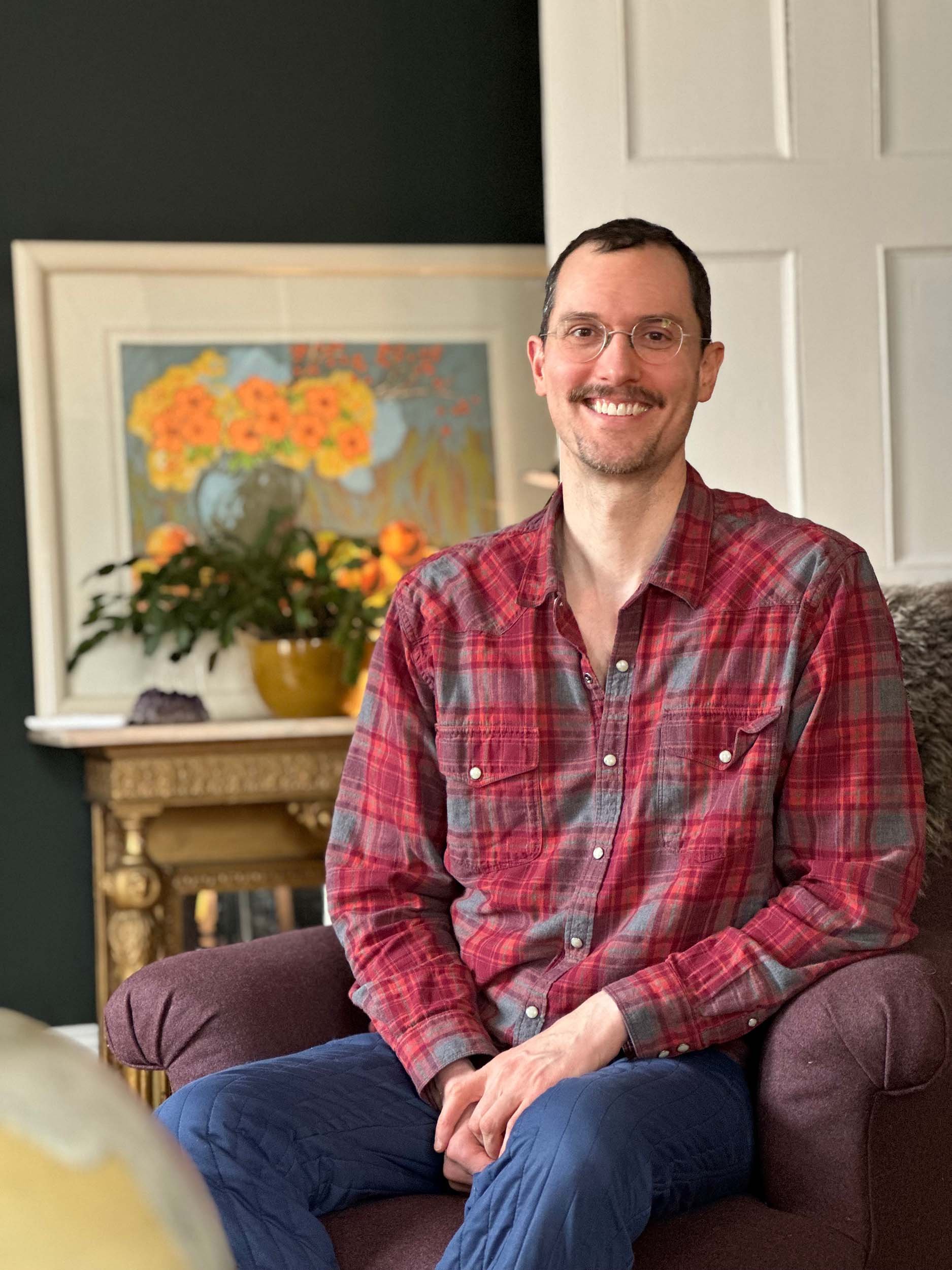 A man with glasses, a beard, and a smile wearing a red plaid shirt sitting on a purple chair in a cozy living room with artwork, a plant, and a wooden table in the background.