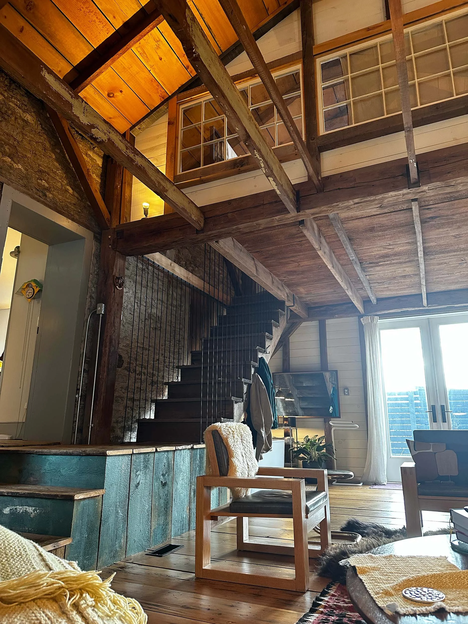 Living room with rustic wooden ceiling and walls, featuring a staircase with black steps and a wooden railing, a TV, large window with cream curtains, and various furniture and decorative items.