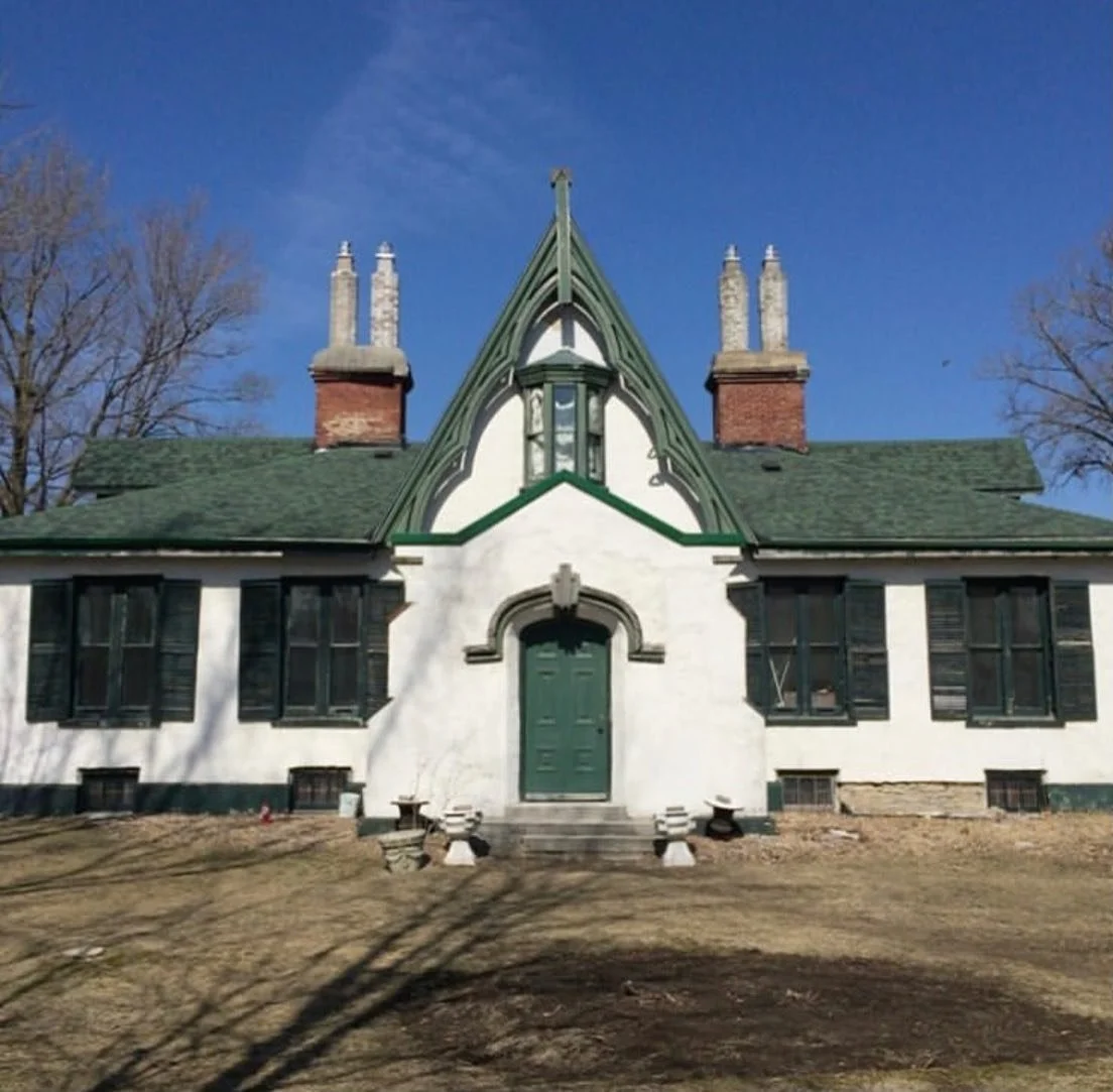 Front view of a historic house with Tudor-style architecture, white stucco walls, dark green trim, a green door, and multiple chimney stacks, set against a blue sky with leafless trees.