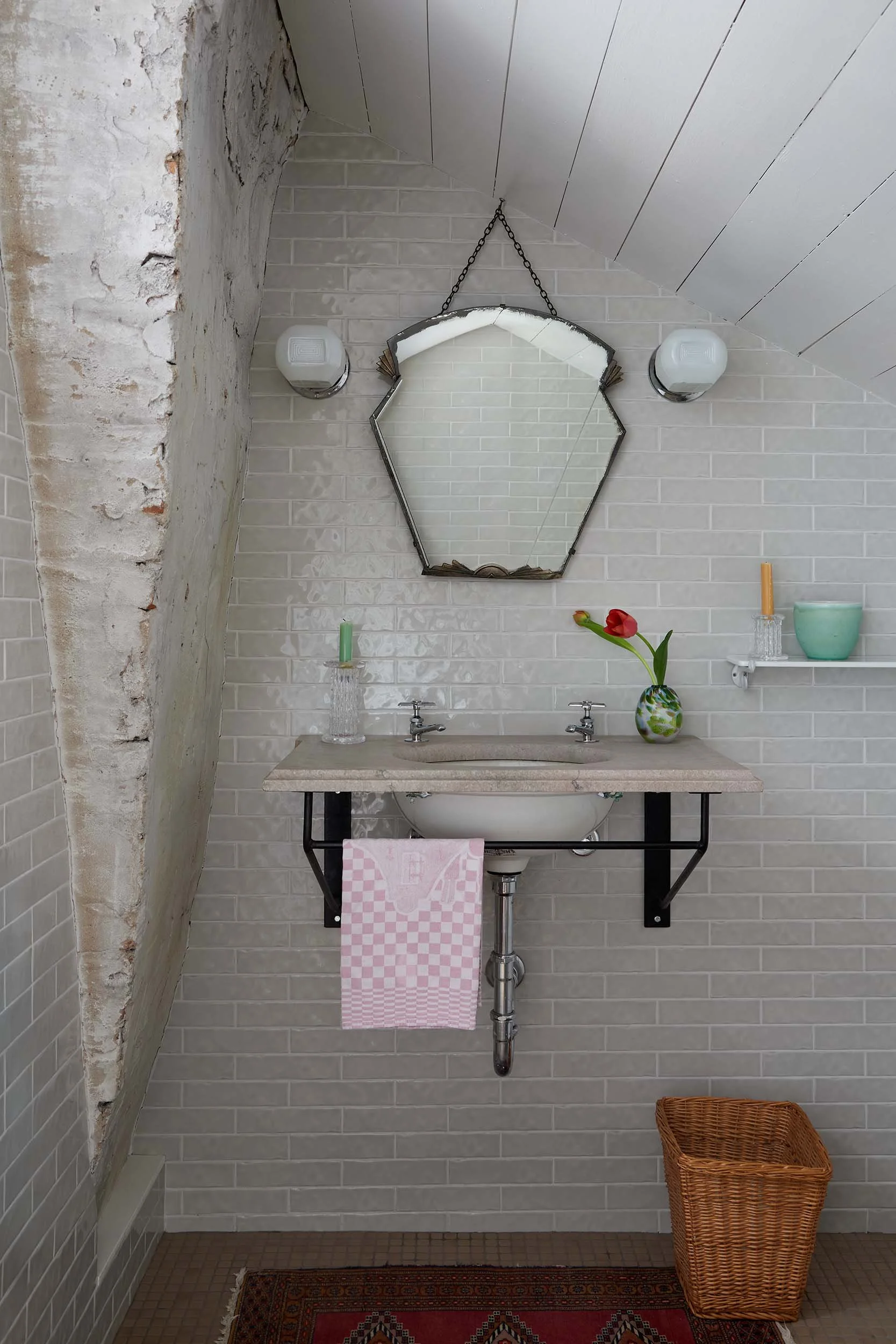 Bathroom sink area with a beige countertop, a sink faucet, a pink checkered towel hanging, a wicker laundry basket, a colorful rug, a flower in a vase, wall-mounted lights, a mirror, and visible brick repairs on the upper corner of the wall.