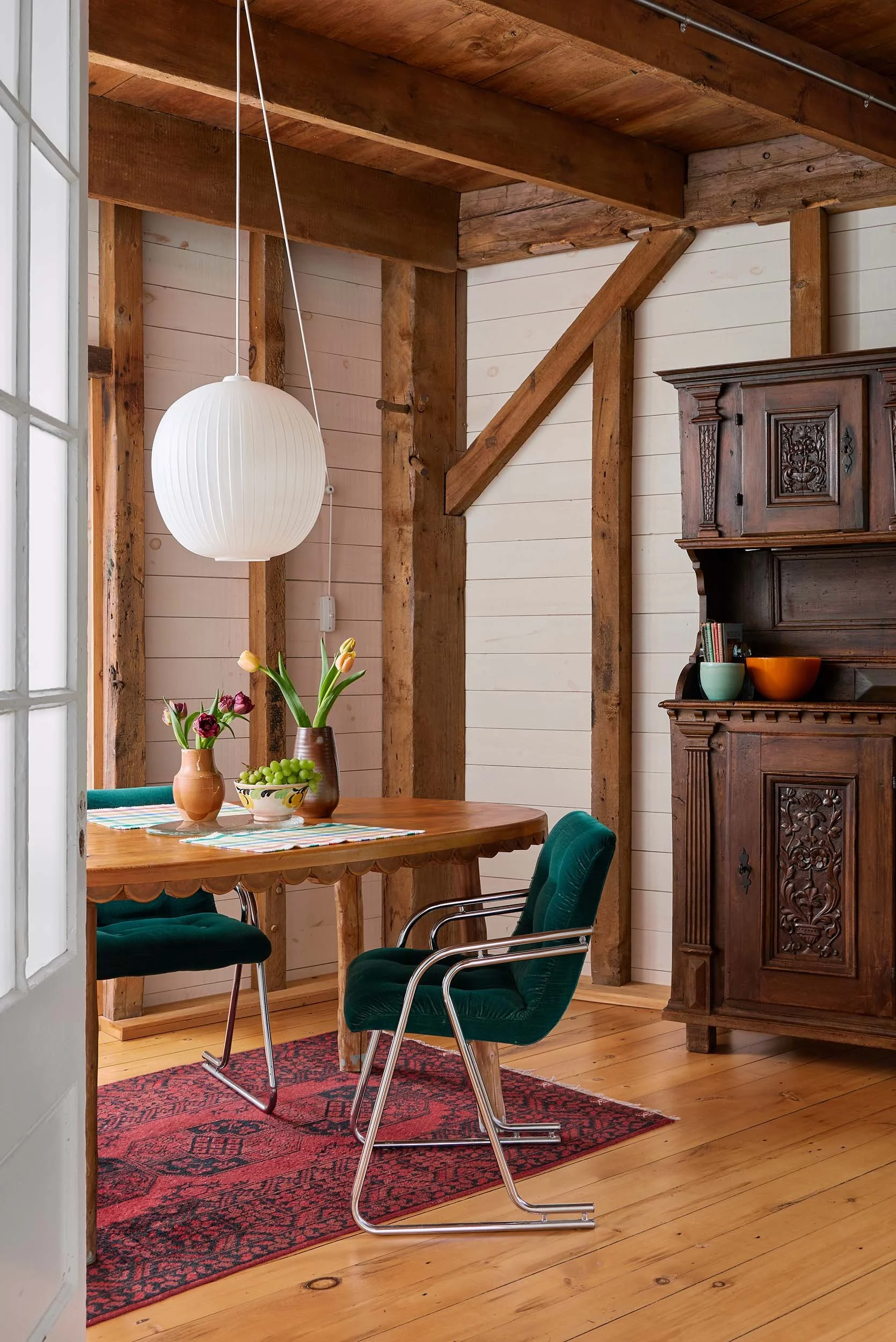 Dining room with a wooden table, green chairs, a red patterned rug, a wooden cabinet, a white paper lantern pendant light, a vase with tulips, a bowl of grapes, and a window with natural light.
