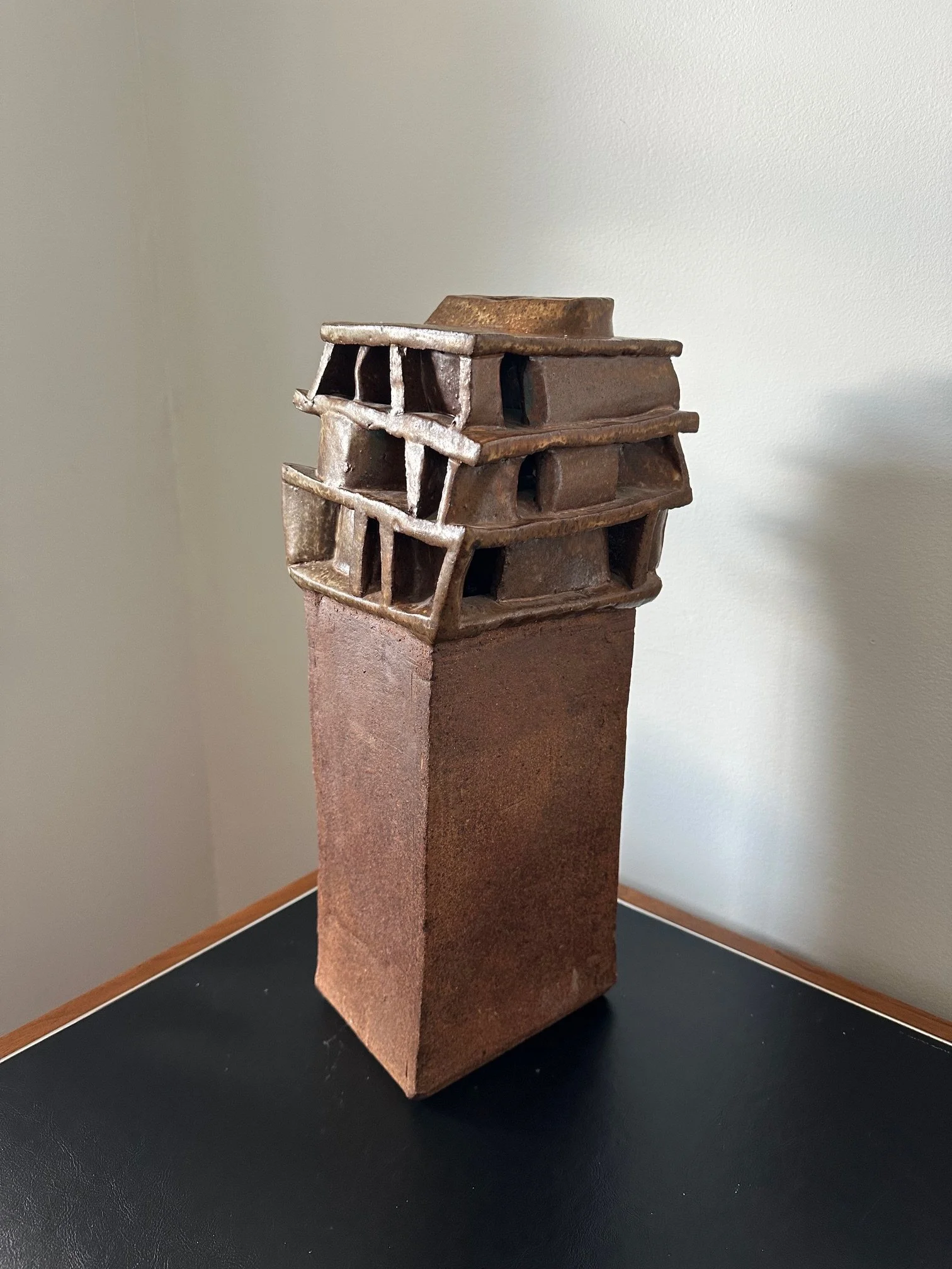 A small sculpture of a multi-story building, made of metal, sitting on a tall, rectangular, brown pedestal with a black surface.