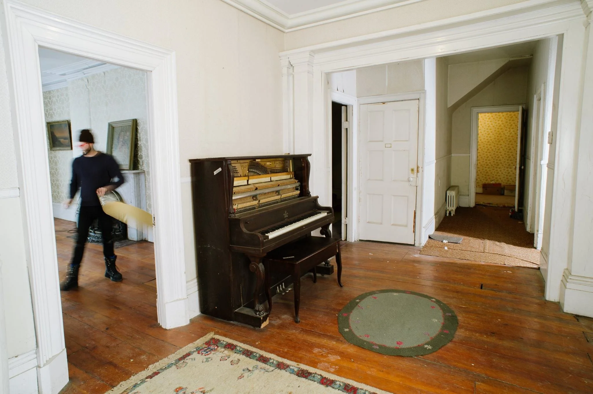 Interior of an old house with wooden floors, a vintage piano, and patterned rugs. A person wearing black clothing and boots is walking through the room, which has white walls and door frames, and visible doorways leading to different rooms.