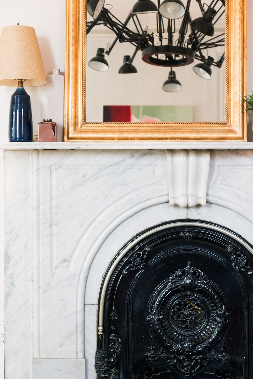 Mirror reflecting a ceiling chandelier with multiple black spotlights, placed above a fireplace with a white marble mantel and ornate black fire grate. On the mantel, there is a beige lampshade with a dark blue base, a pink box, and a partial view of a green plant.