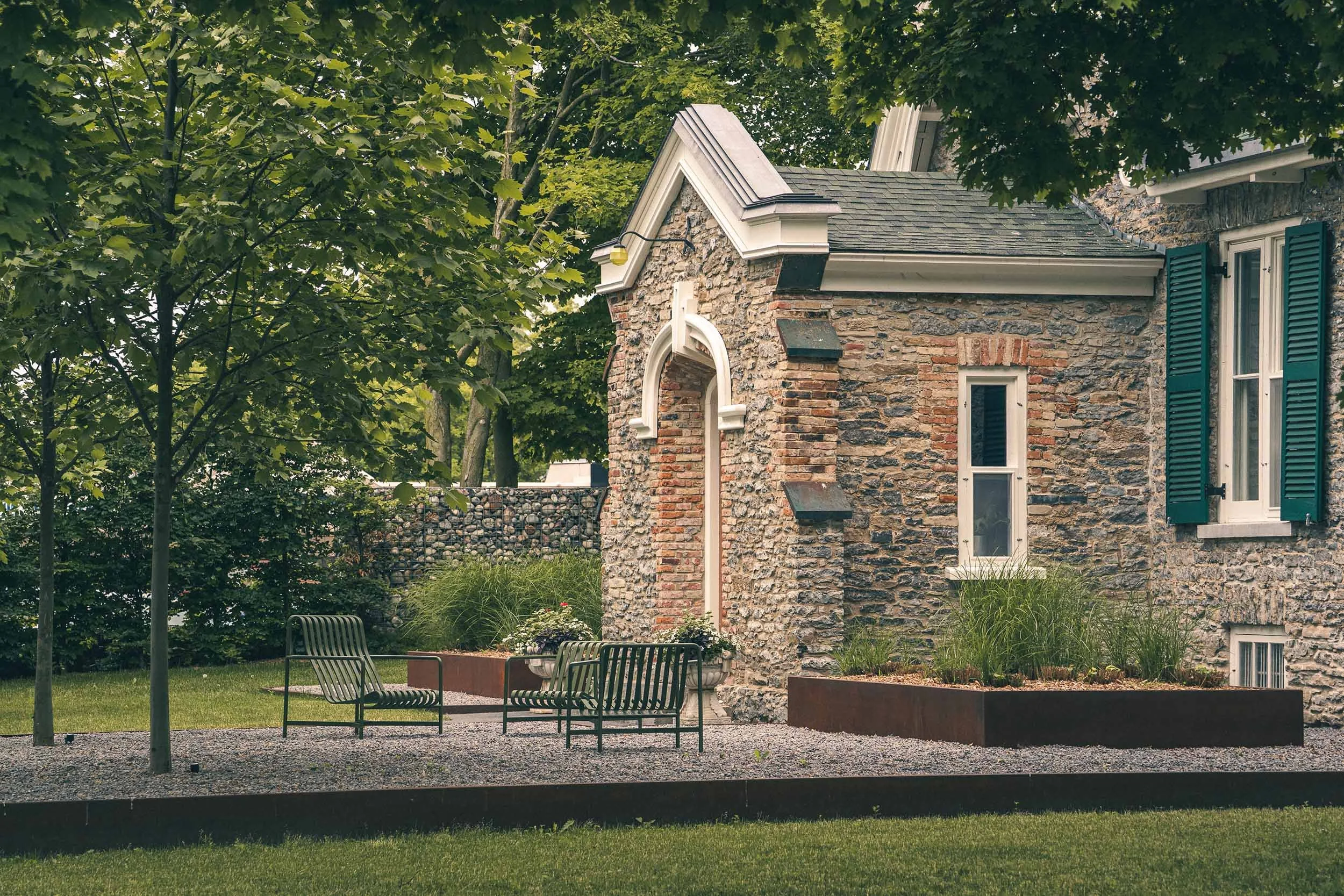 A house with stone exterior walls, white window shutters, and a steep gabled roof, surrounded by green trees and a garden area with metal chairs.