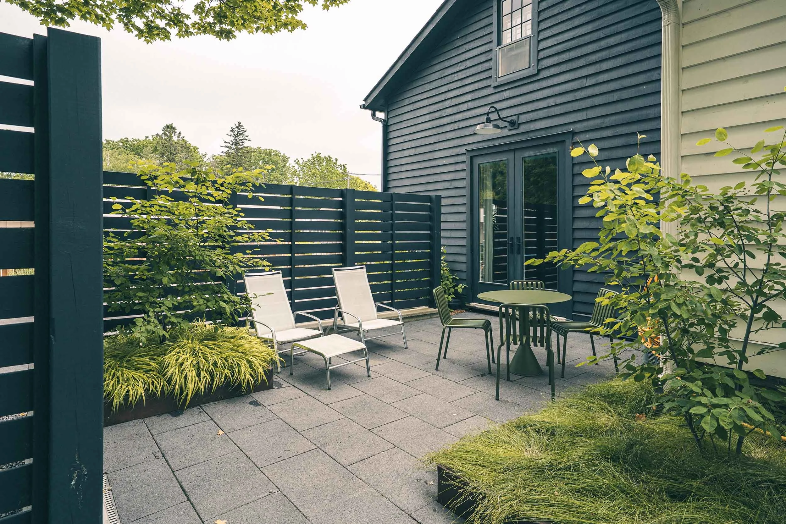 A backyard patio with outdoor chairs, tables, and lush plants, enclosed by a dark blue wooden fence, adjacent to a dark blue house with a sliding glass door and exterior lighting.