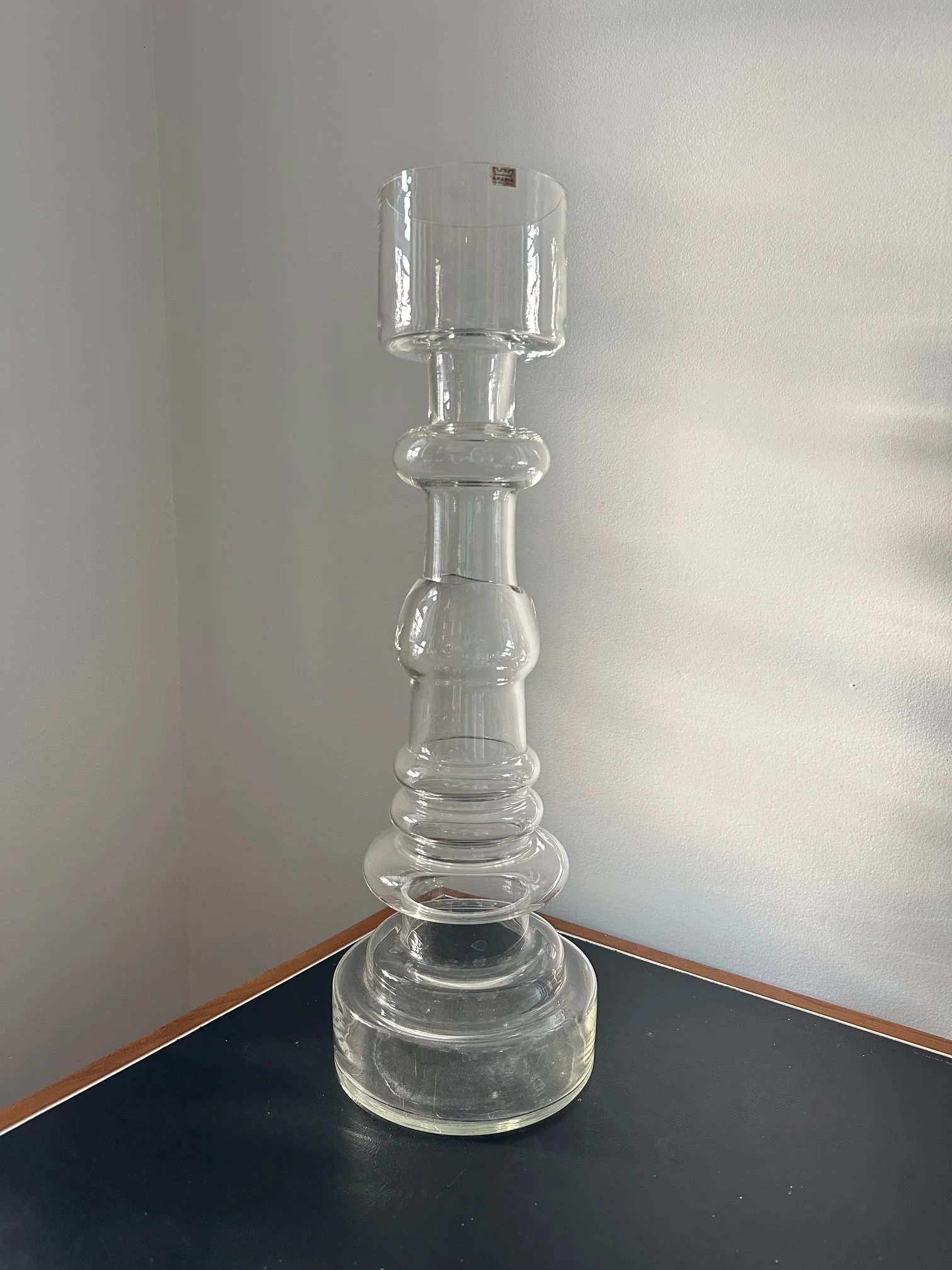 A tall, tiered glass sculpture with multiple interconnected transparent glass segments arranged vertically, placed on a black surface against a light-colored wall.