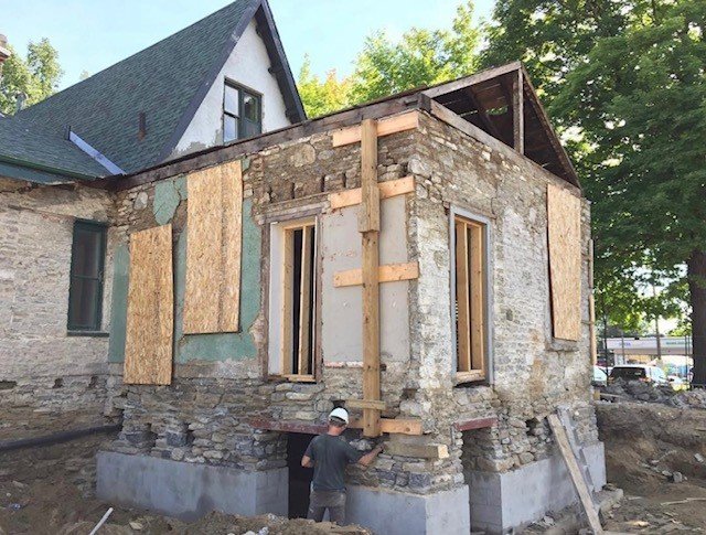 Construction worker working on the foundation of a house under renovation, with new brickwork and plywood panels visible on the exterior walls.