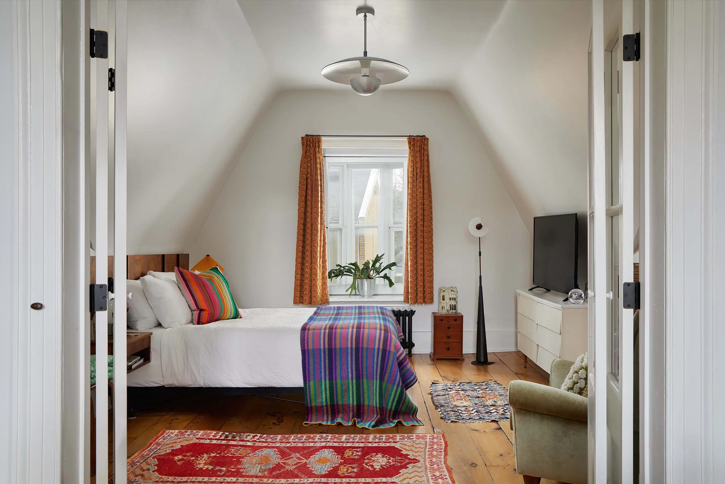 A cozy bedroom with a large bed with white bedding and colorful striped pillows. A window with orange curtains and a plant on the sill. A television on a white dresser, a floor lamp, and a small table with a decorative object. The room has a sloped ceiling and wooden floor.