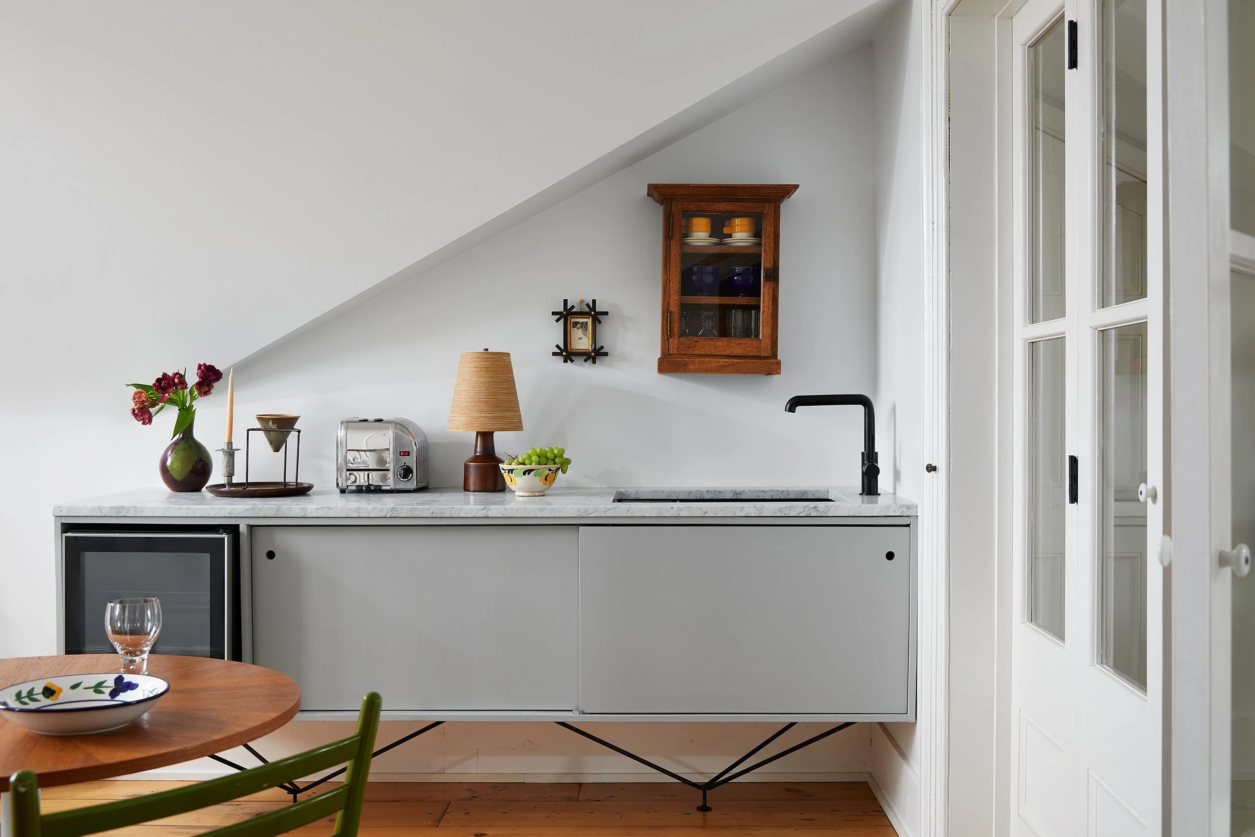 A kitchen with a white countertop, a small wooden table with a floral plate and green chair, a vase with flowers, a toaster, a lamp, a bowl of grapes, a black faucet, a wooden cabinet with glass doors mounted on the wall, and white paneled walls and doors.