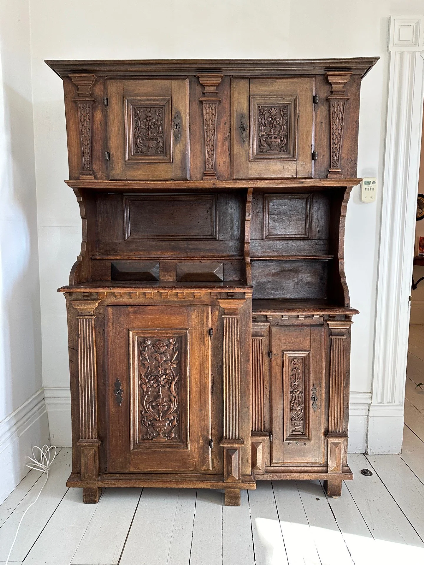 An antique wooden cabinet with ornate carved details, standing against a light-colored wall inside a room with a white floor.