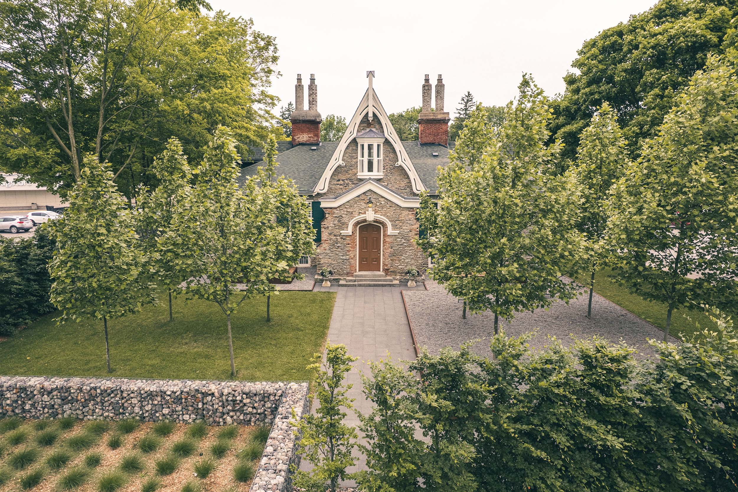 A stone house with a steep gabled roof, surrounded by green trees, with a pathway leading to the front door and a well-maintained yard.