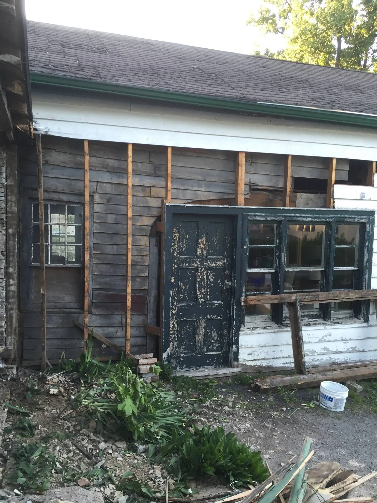 The image shows the side of a house that is under renovation. The exterior wood siding is partially removed, exposing vertical wooden studs. There is a black, damaged door leaning against the house, and some construction debris, including a white bucket, in the foreground. Green plants and dark soil are also visible at the bottom of the image.