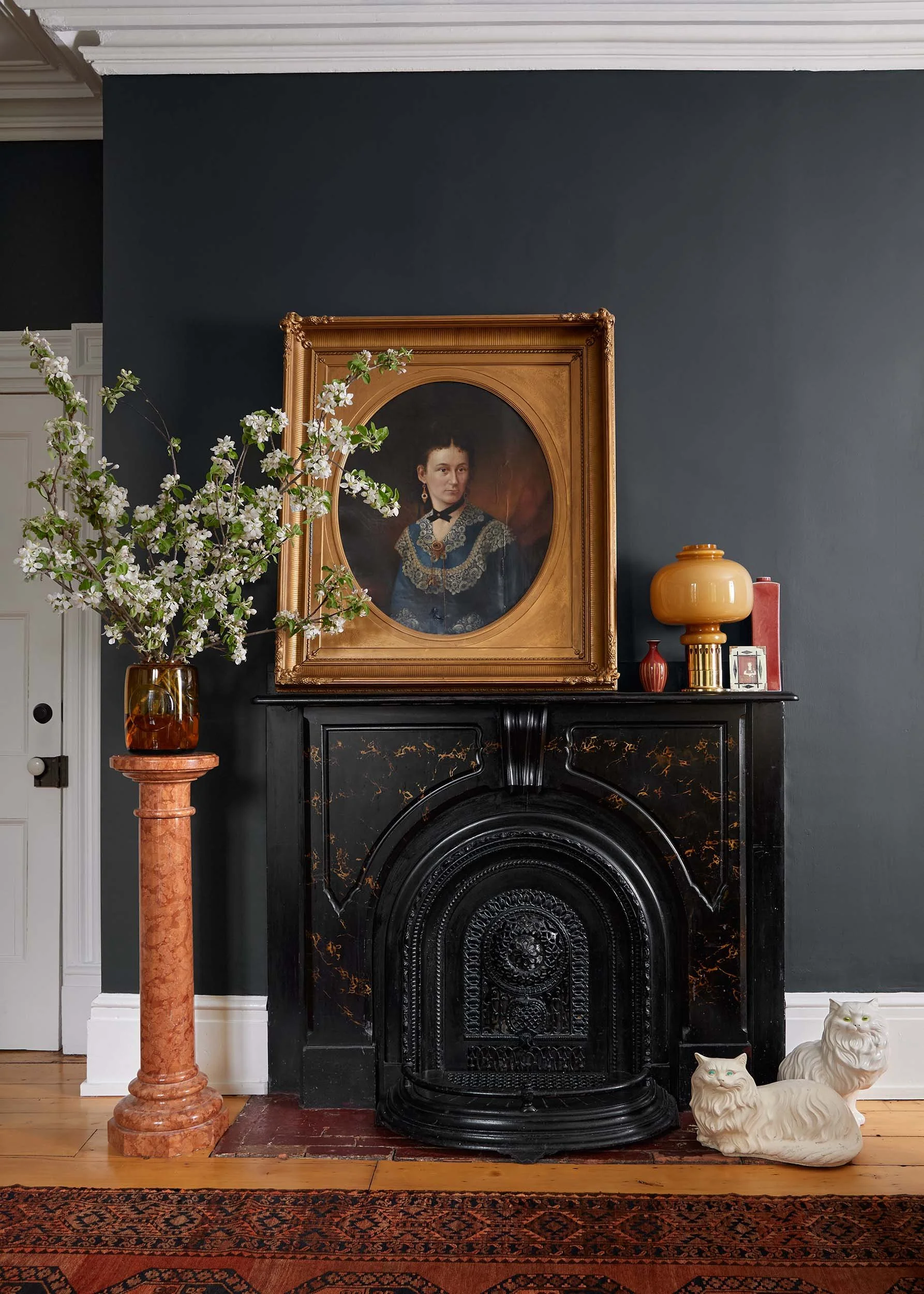 Interior of a room featuring a black fireplace with ornate detailing, a framed portrait of a woman in vintage attire above the fireplace, a large vase with white flowering branches on a pink marble pedestal to the left, and three ceramic lion figurines on the floor to the right, with a dark wall behind.