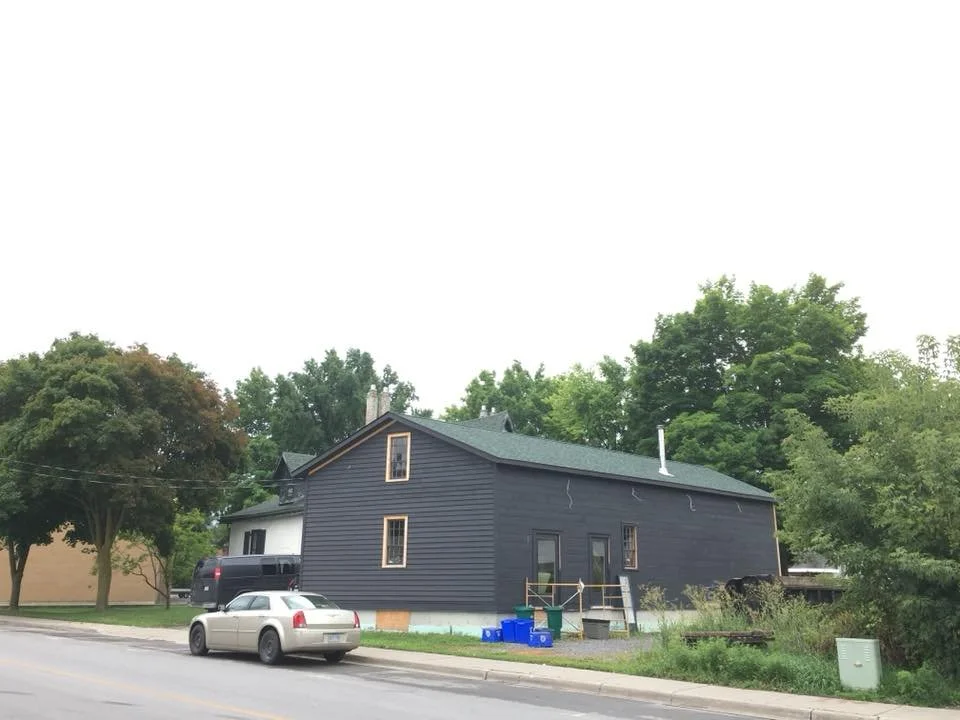 A two-story house under construction with black siding, a green roof, and a small set of stairs leading to a door. A white car and a black truck are parked in front, with construction materials and blue containers nearby. There are large trees and other buildings in the background.