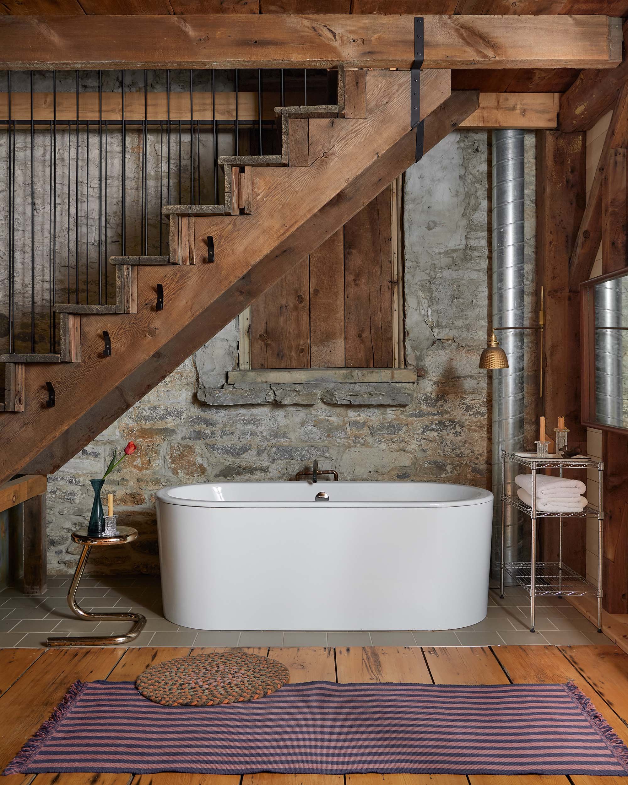 A rustic bathroom with a white freestanding bathtub placed on a tile floor. The bathtub is set against a weathered stone wall with unfinished wooden paneling. There is a small round side table with a glass and a flower vase next to the tub, and a metal shelving unit with rolled towels on the right. The ceiling and support beams are made of natural wood, and there are decorative rugs on the wooden floor in front of the tub.