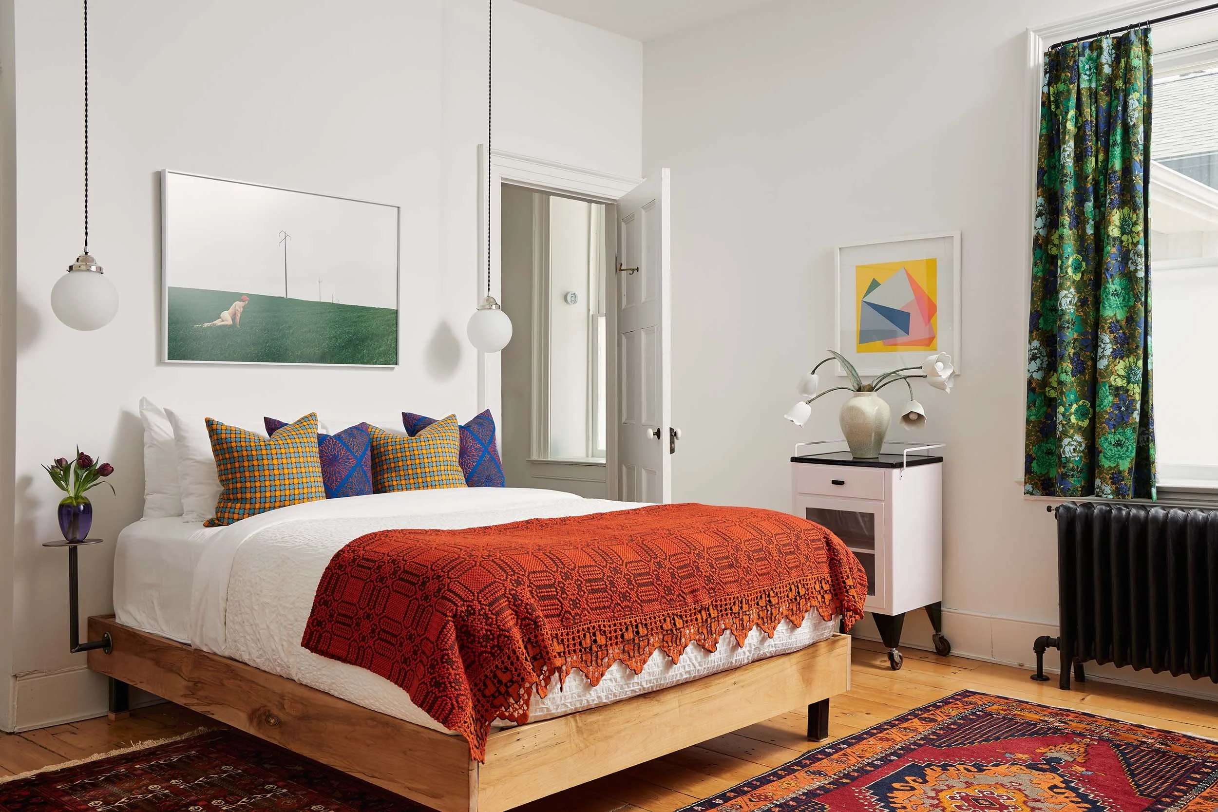 A cozy bedroom with a wooden bed, colorful pillows, white bedding, a red throw blanket, and a side table with a purple vase and flowers. There are hanging globe lights, artwork on the walls, a window with floral curtains, and a vase with flowers on a white dresser.