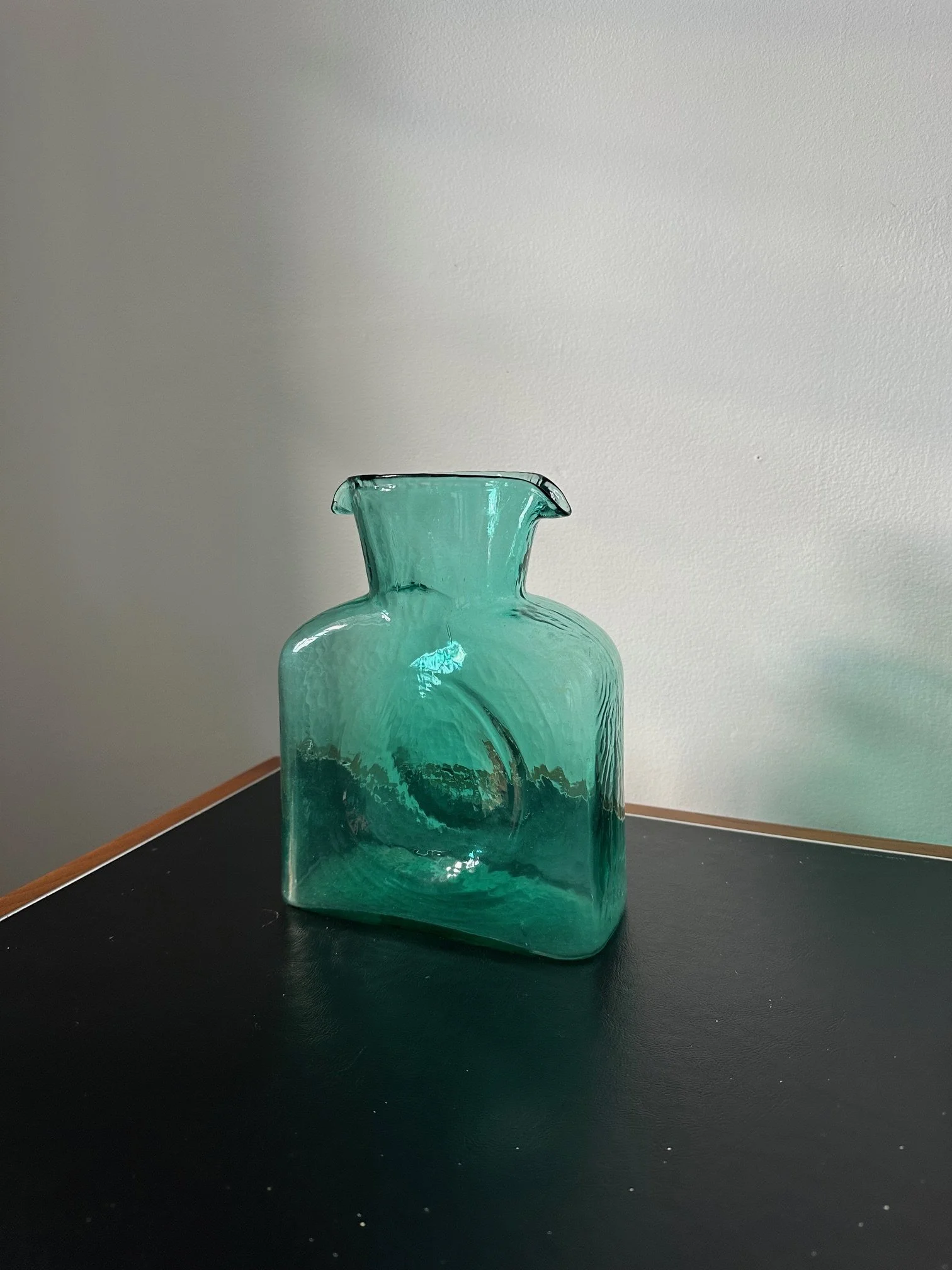 A teal-colored glass vase with a wide, flared open top and a rectangular body, placed on a black surface against a light-colored wall.