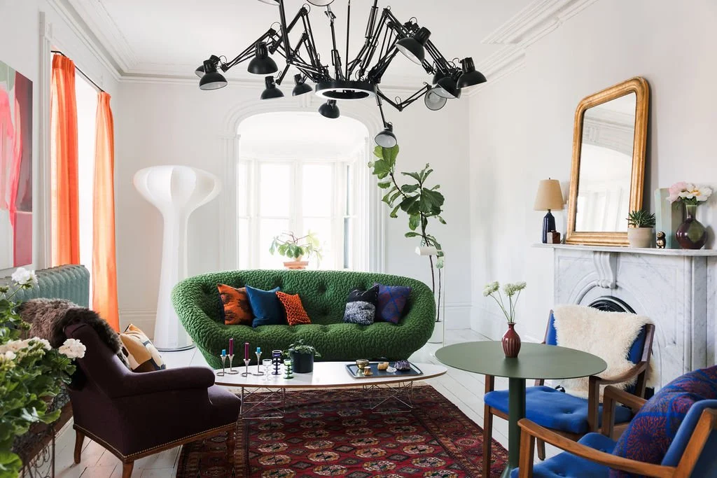 Bright living room with white walls, large windows, orange curtains, and a unique black chandelier. Features a green textured sofa with colorful pillows, a dark purple armchair with patterned cushions, a small round gray table with a red vase and white flowers, and a decorative white sculpture near the window. A large mirror is above the white fireplace mantle decorated with lamps, plants, and vases. The room has a colorful patterned rug and a white floor lamp.