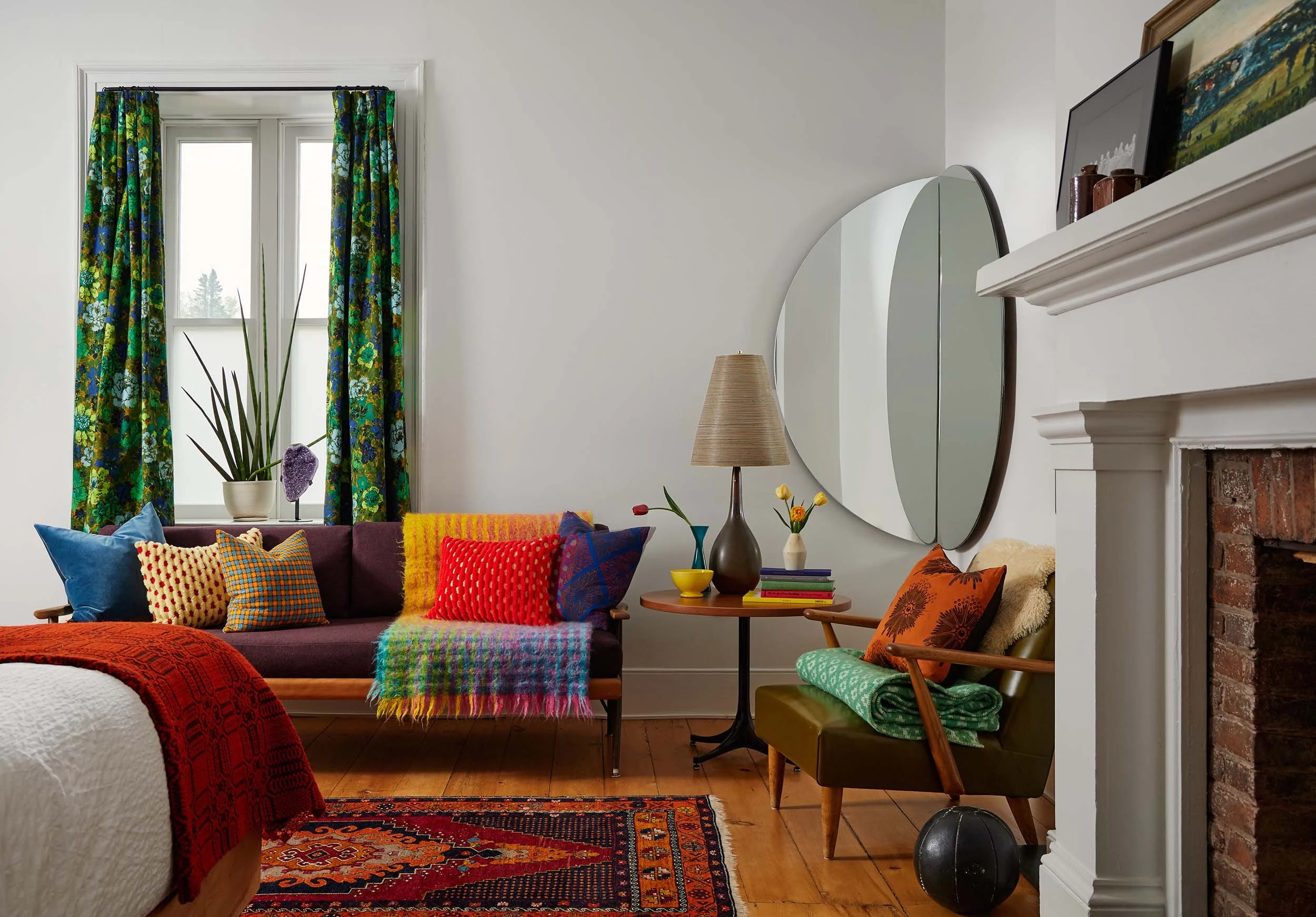 A cozy living room with colorful cushions on a couch, a rainbow plaid blanket, a plant on a window sill, and a brick fireplace. The room features a large, round mirror, a table with decorative vases, a floor lamp, and vibrant curtains with a floral pattern.