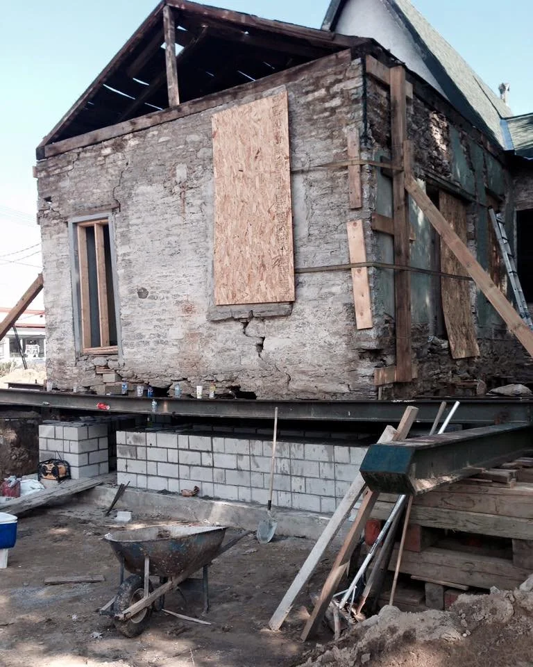 A house under construction with wooden framing, plywood panels, and brick foundation blocks, along with various construction tools and materials.