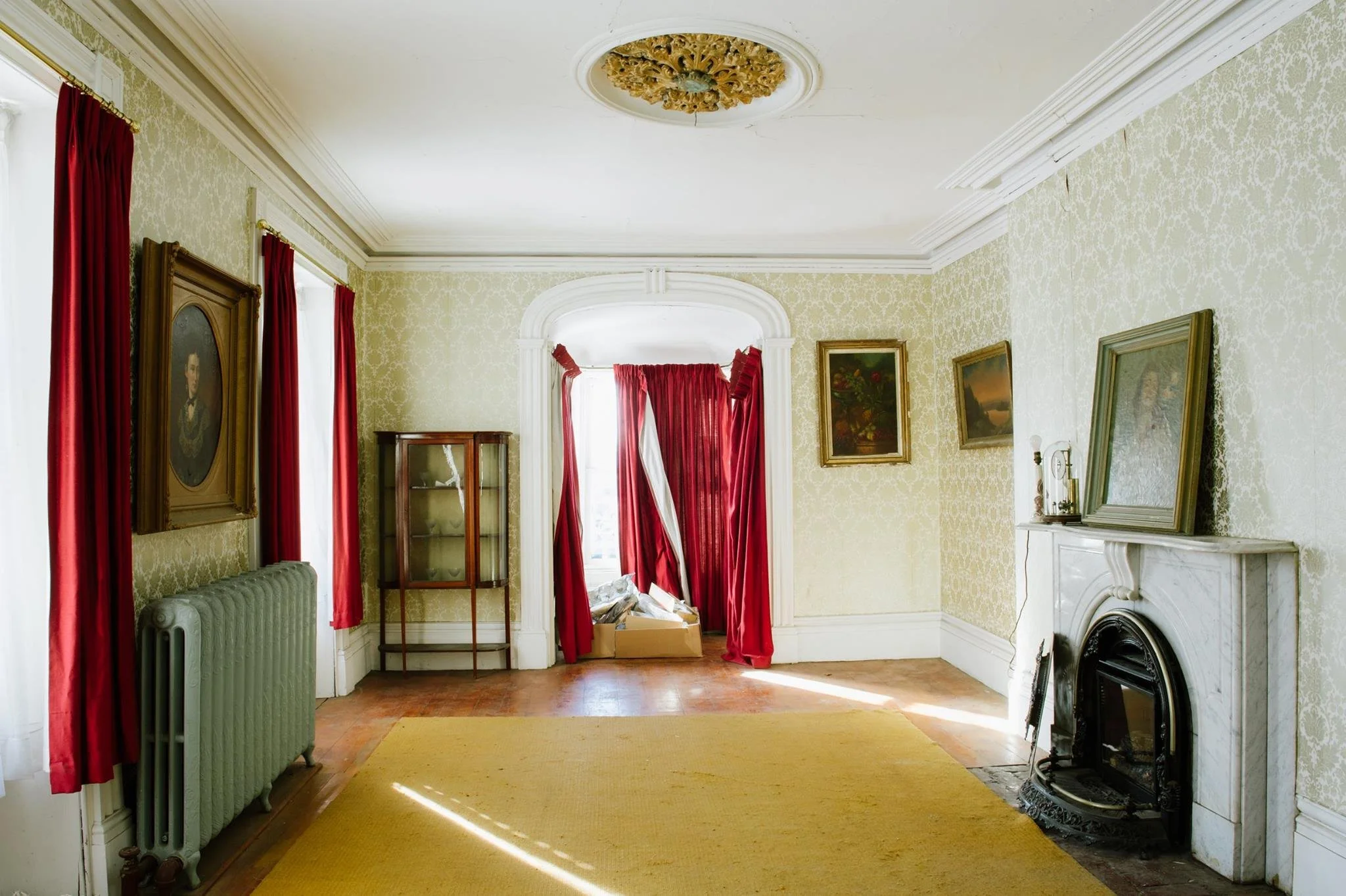 Living room with ornate ceiling medallion, large windows with red curtains, framed artwork, and a fireplace, in a vintage style.