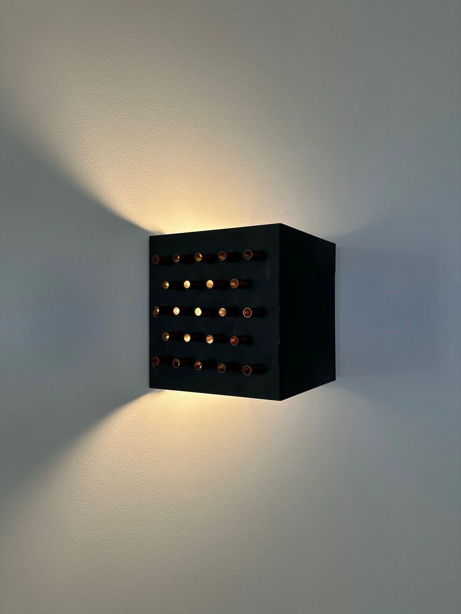 A black wall-mounted wine rack with circular holes, illuminated by a light source on a white ceiling and wall.