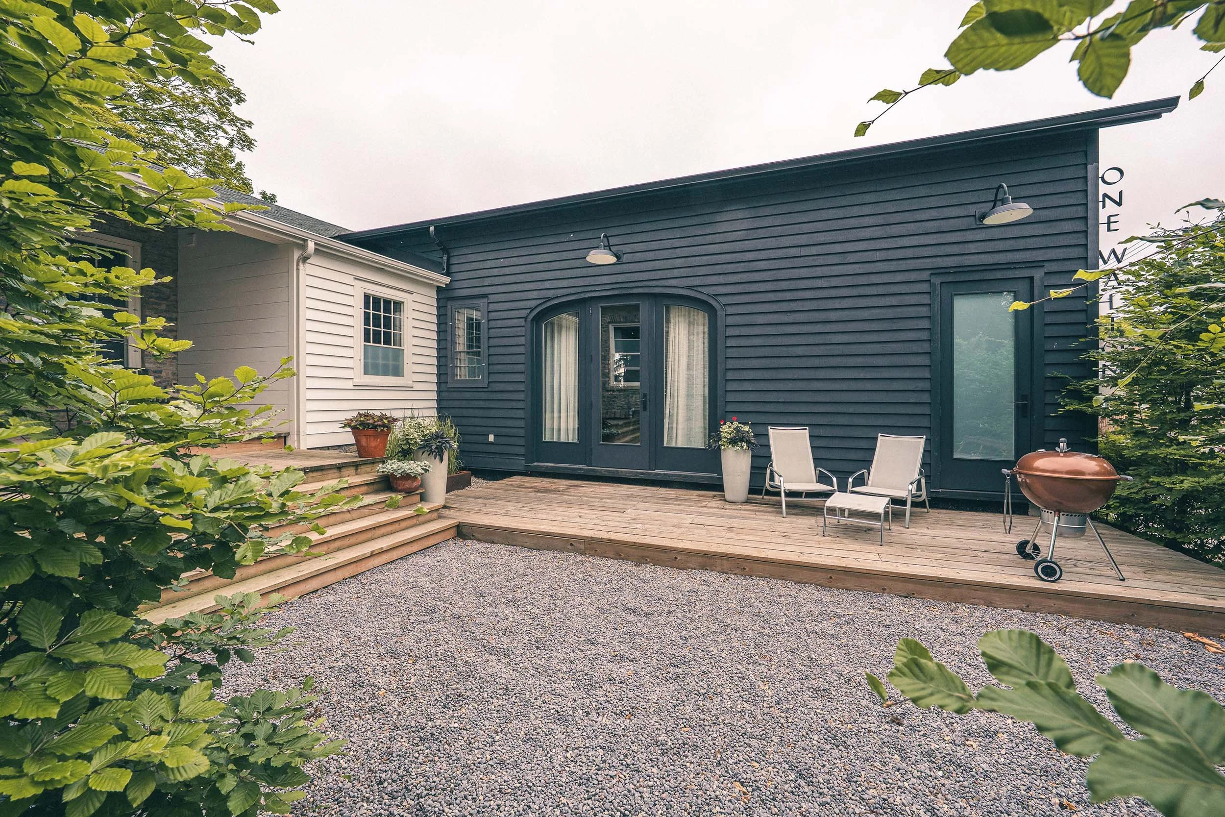 Backyard patio with wooden deck, outdoor chairs, potted plants, and a charcoal grill, surrounded by green foliage and trees, with a modern house featuring a black and white exterior.
