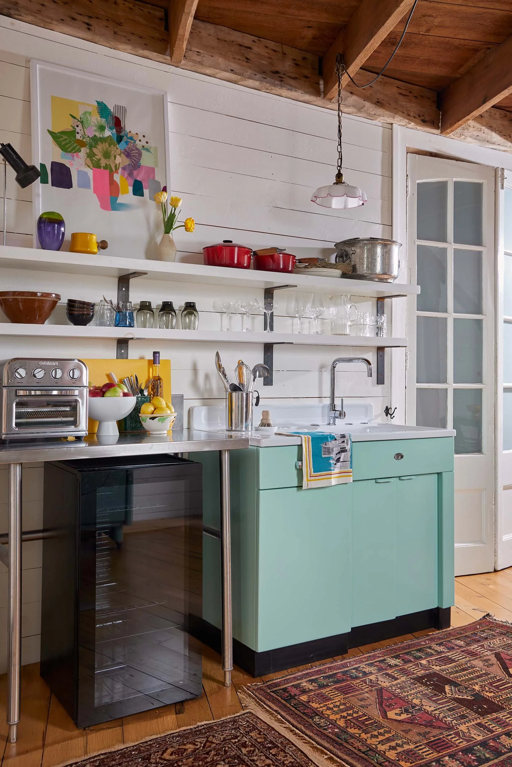 A kitchen with white paneled walls, a turquoise lower cabinet, and open shelves holding various dishes and glassware. There is a black microwave on the floor, a small stainless steel table with a bowl of apples, and a black oven. The countertop has a fruit bowl, some utensils, and a towel hanging from the cabinet handle. A rug with a traditional pattern covers part of the wooden floor, and a colorful framed floral artwork hangs on the wall. There is a window with a white frame and a vintage hanging light fixture.