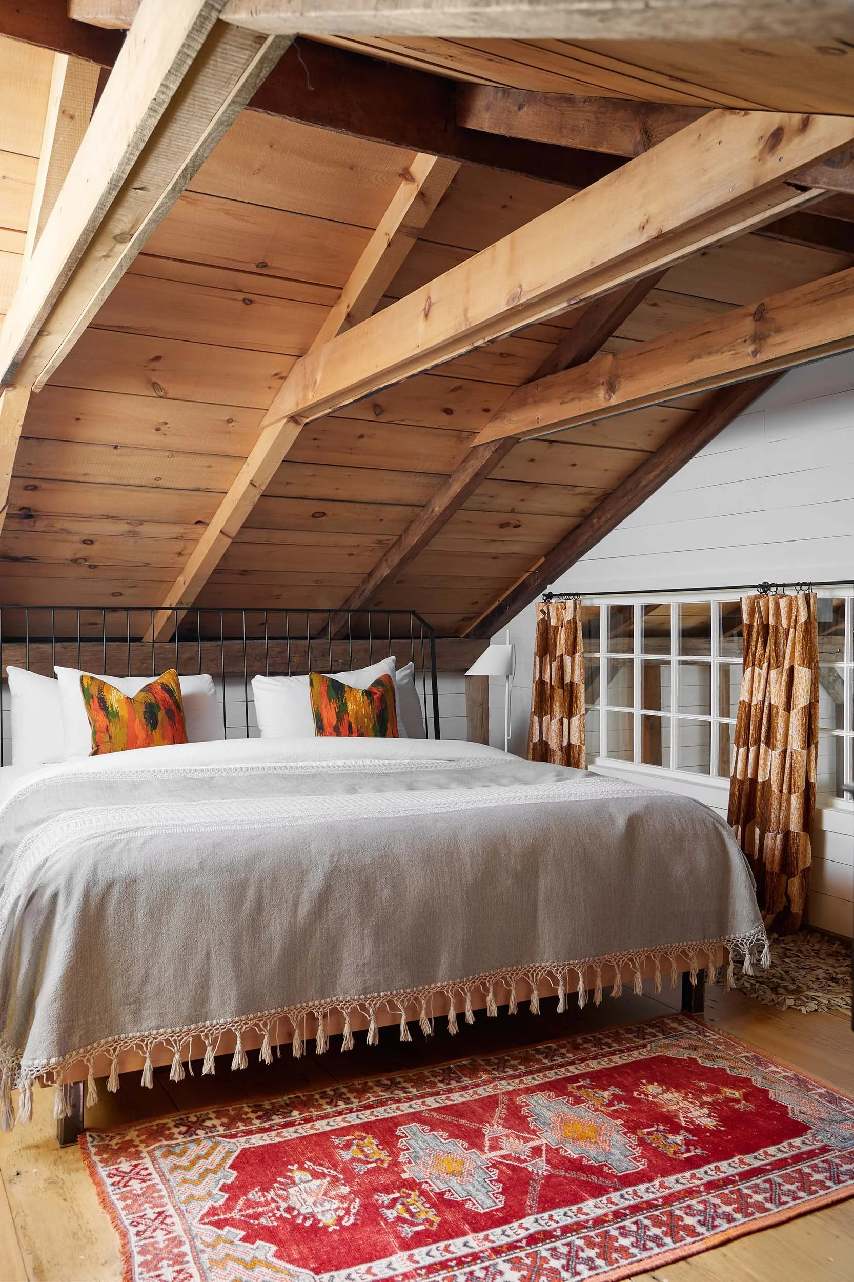 A cozy bedroom with a bed dressed in white bedding and colorful pillows, under a wooden vaulted ceiling with exposed beams, next to a window with patterned curtains, and a red oriental-style rug on a hardwood floor.