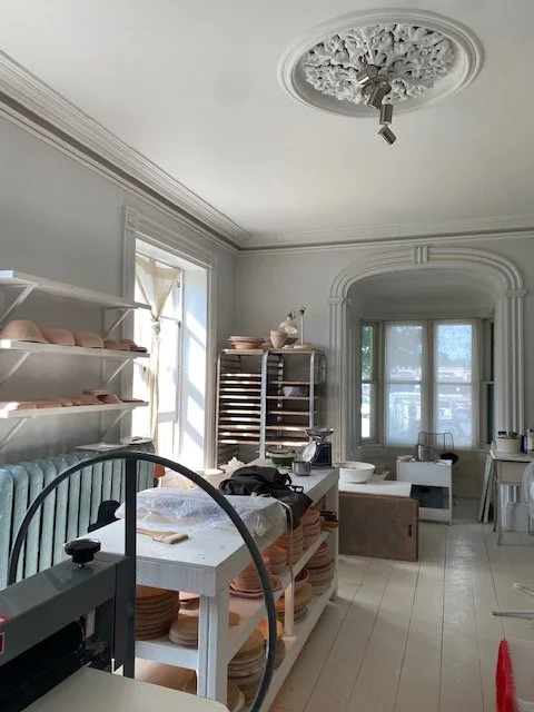 A room with white walls, crown molding, and large windows. There are shelves with pink plates, a table with dishes and kitchen items, and a ceiling medallion with an unused light fixture. The room appears to be a kitchen or dining area, with natural light coming in from the windows.