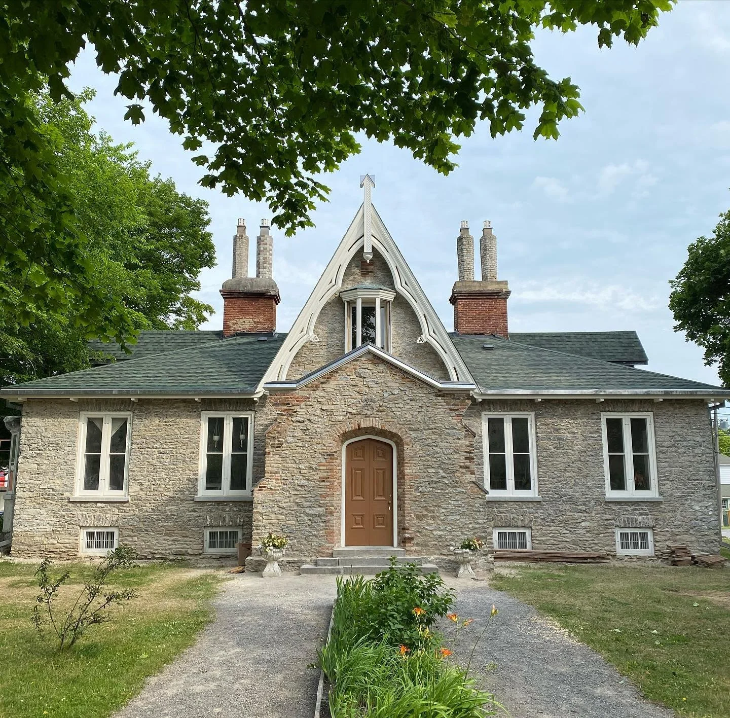 A historic stone house with a steep roof, multiple chimneys, and decorative architectural details, surrounded by greenery and a garden path.