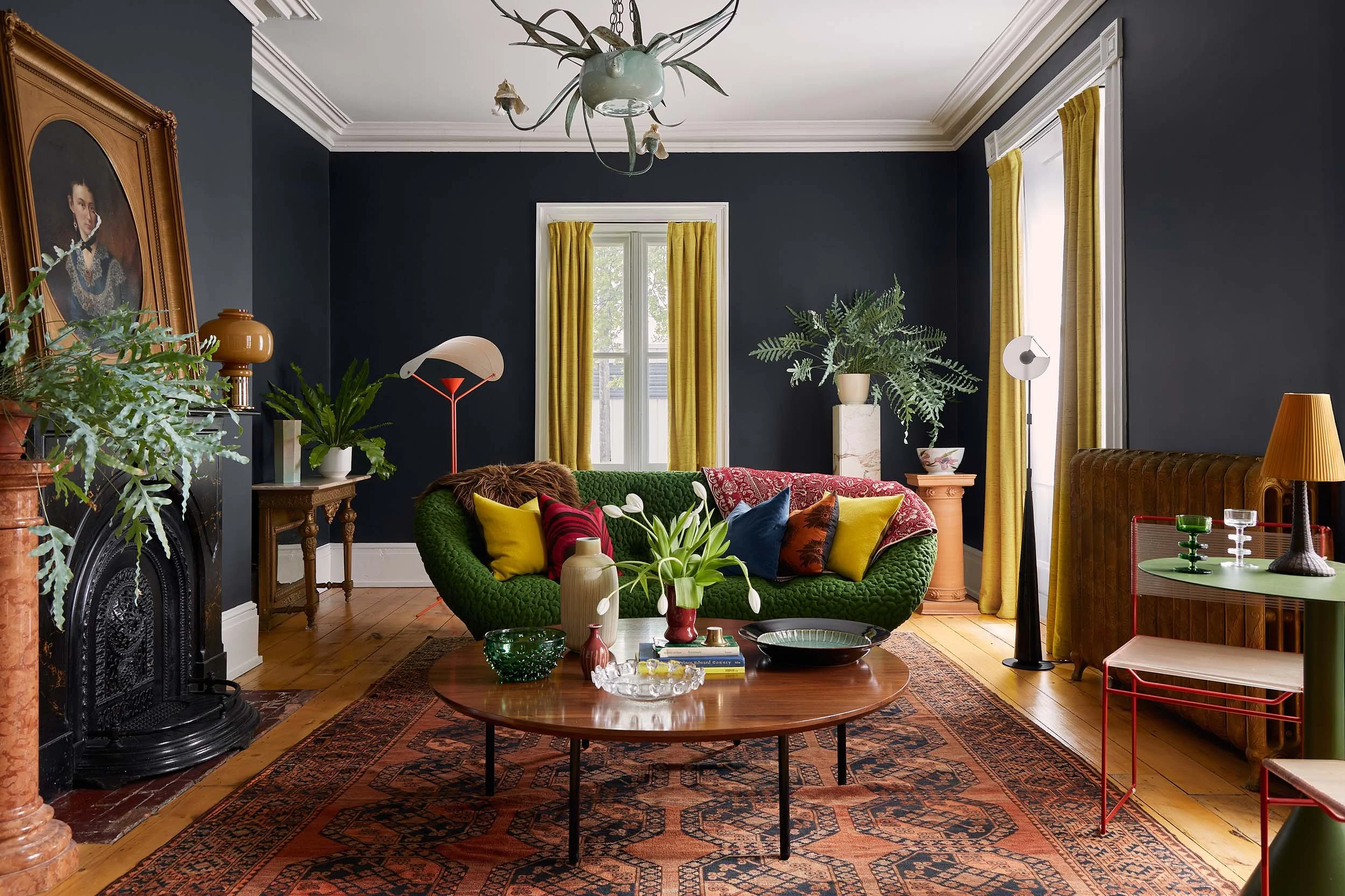 A living room with dark blue walls, yellow curtains, and wooden flooring. Features a green velvet sofa with colorful pillows, a round wooden coffee table with decorative items, and various potted plants. Decor includes an ornate portrait, a fireplace, and modern lighting fixtures.