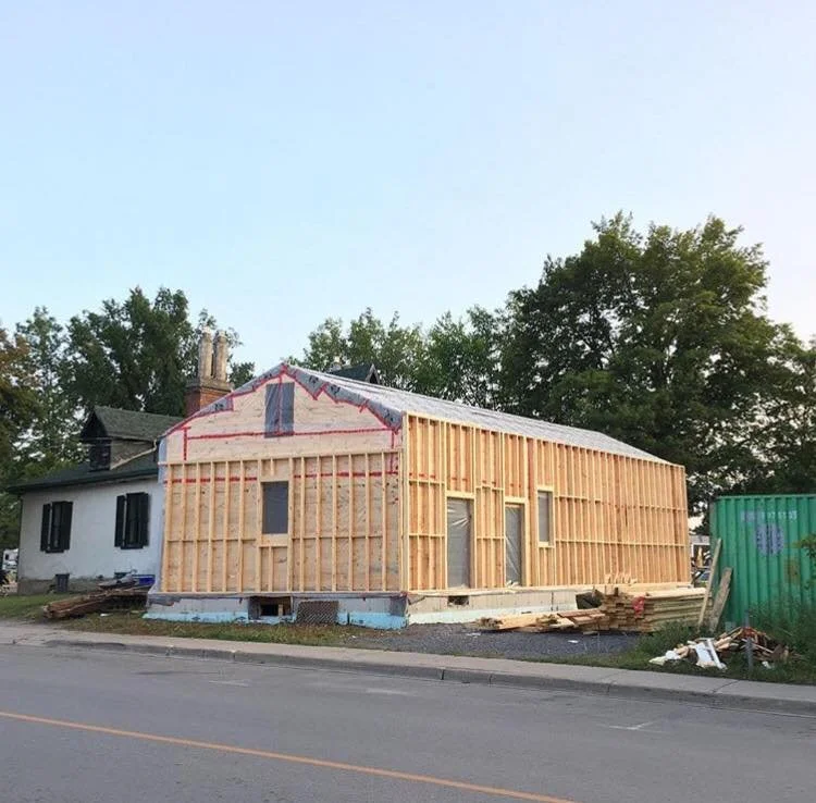 A house under construction, with the exterior walls not yet finished, showing wooden framing and some parts covered with insulation. It is located on a street with an older neighboring house and trees in the background.