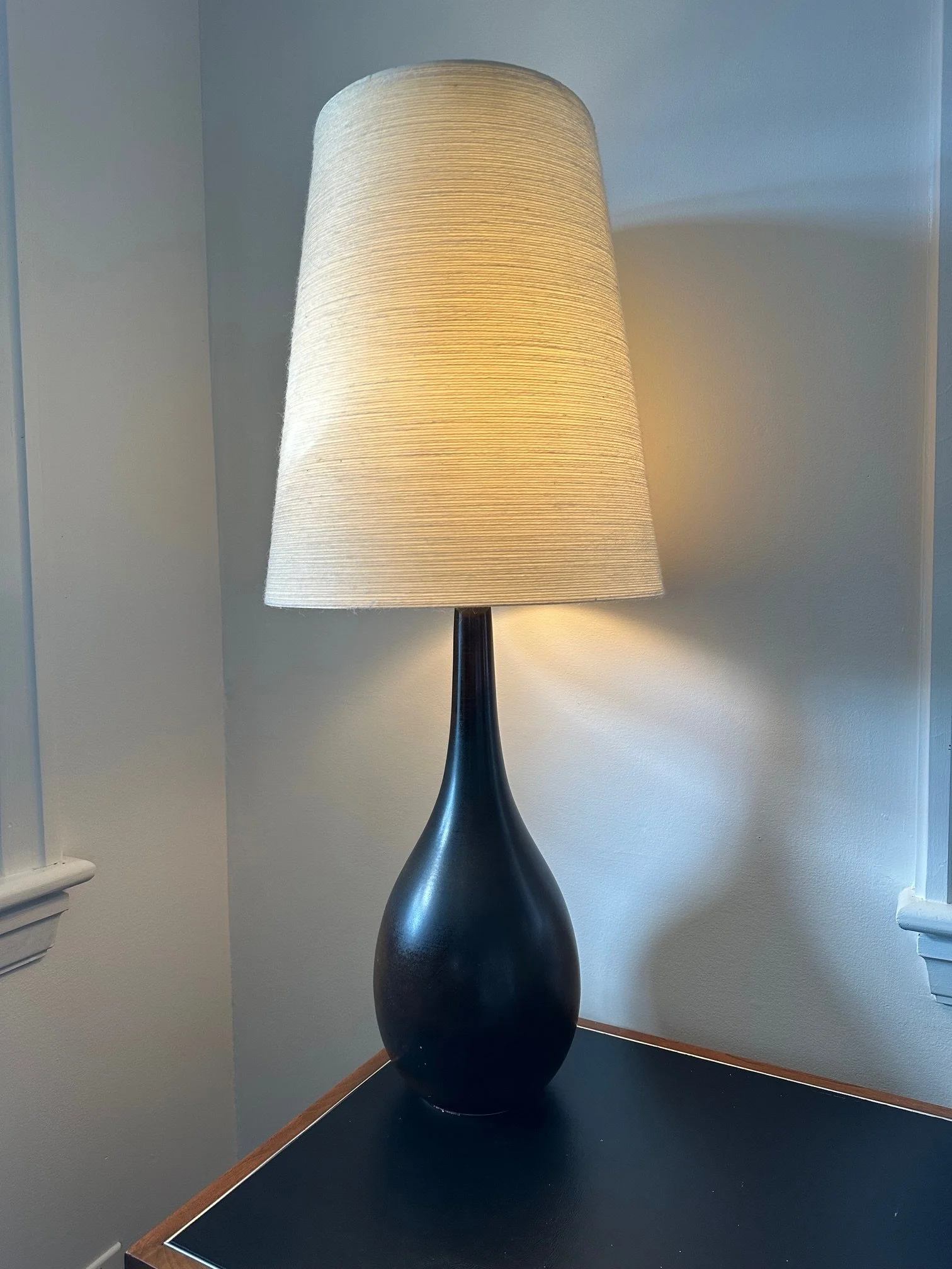 A black ceramic table lamp with a tall beige fabric lampshade, placed on a wooden table against a light-colored wall.