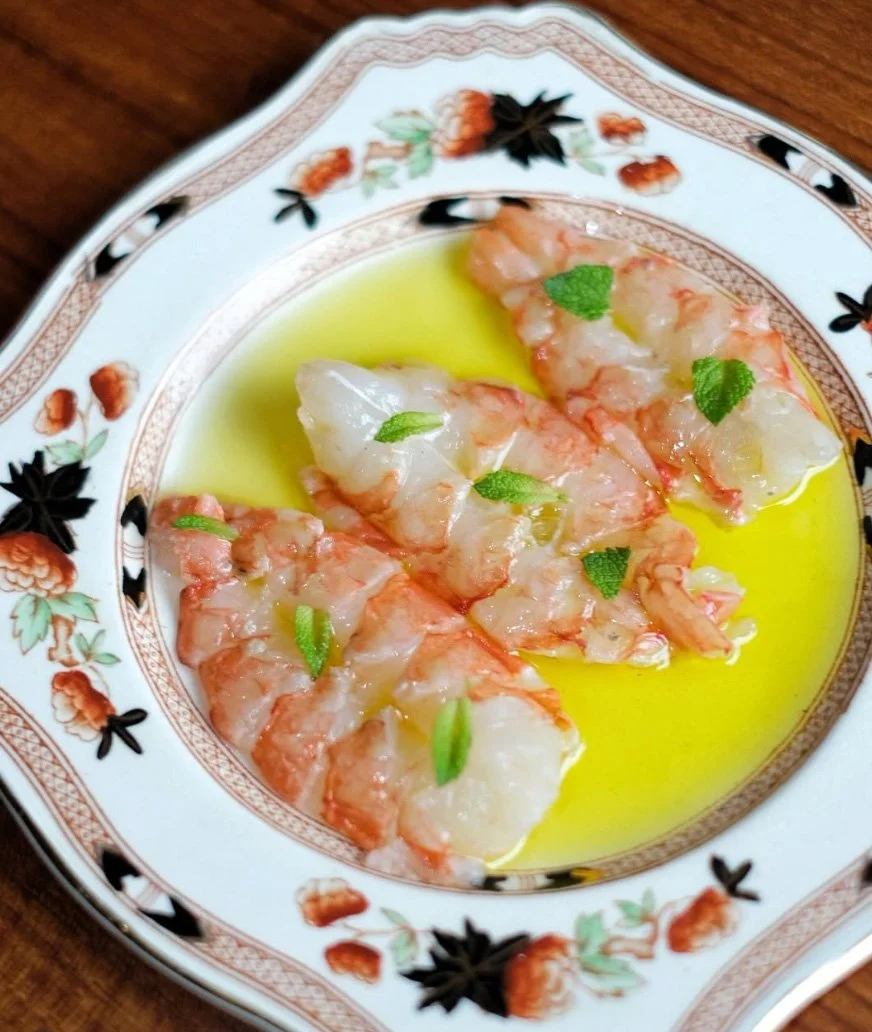 Cooked shrimp topped with green herbs in olive oil on a decorated white and gold floral plate.