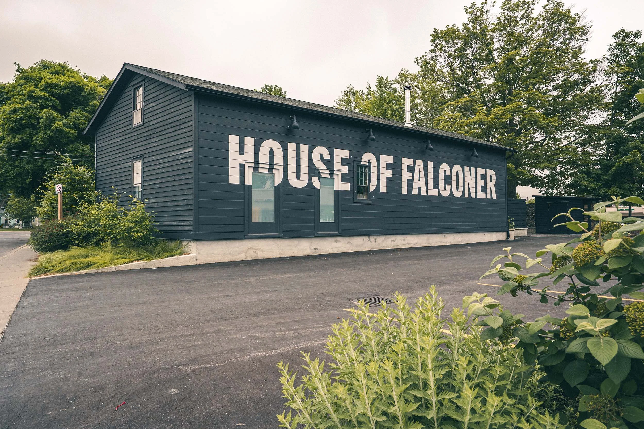 Black building with large white letters reading 'House of Falconer' on its side. Surrounding trees and a driveway in front.