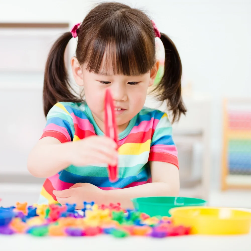 Une jeune fille avec des cheveux en queue de cheval, portant un t-shirt rayé coloré, joue avec des jouets colorés sur une table.